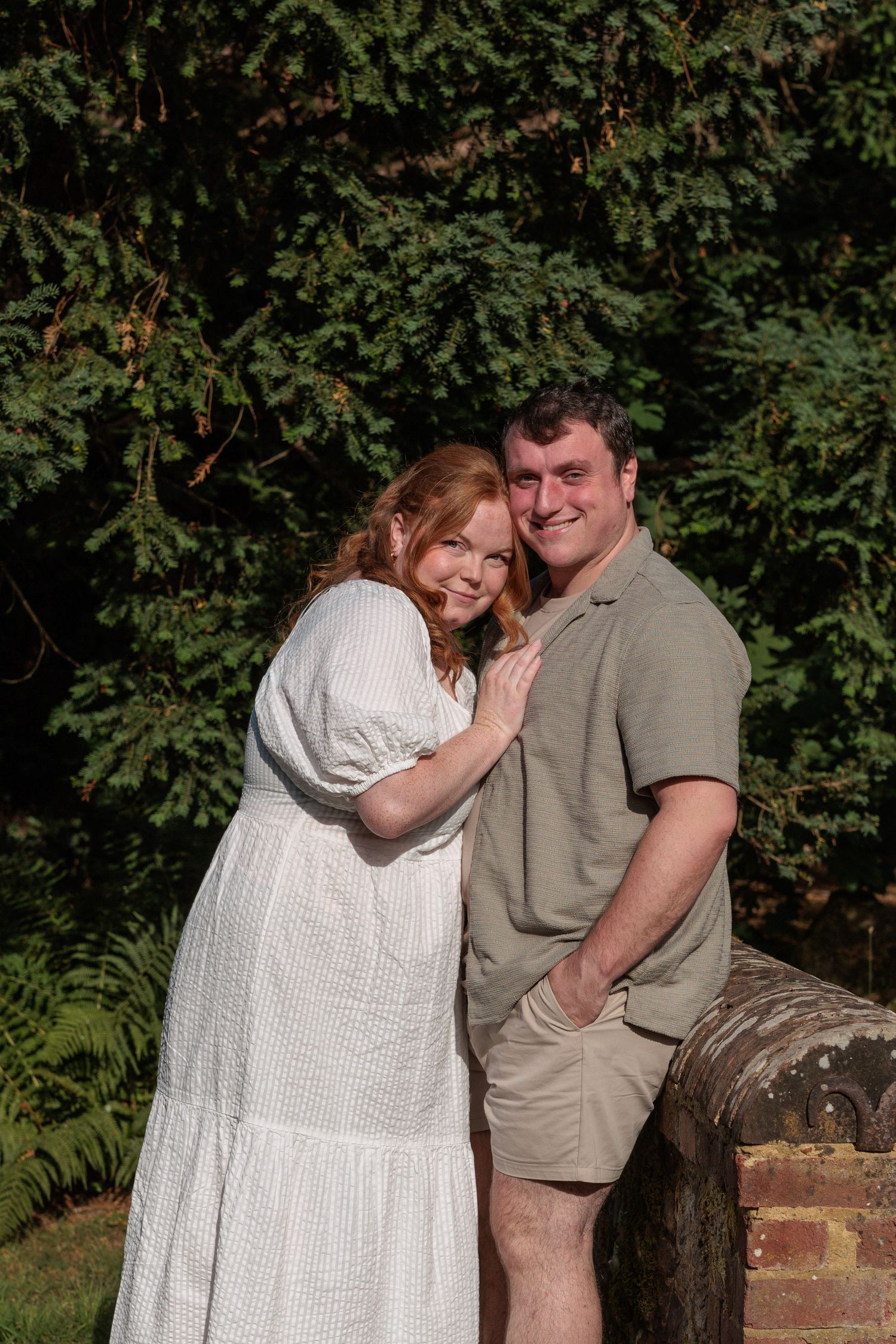 A smiling couple poses outdoors, woman in white dress and man in khaki, surrounded by lush green trees.