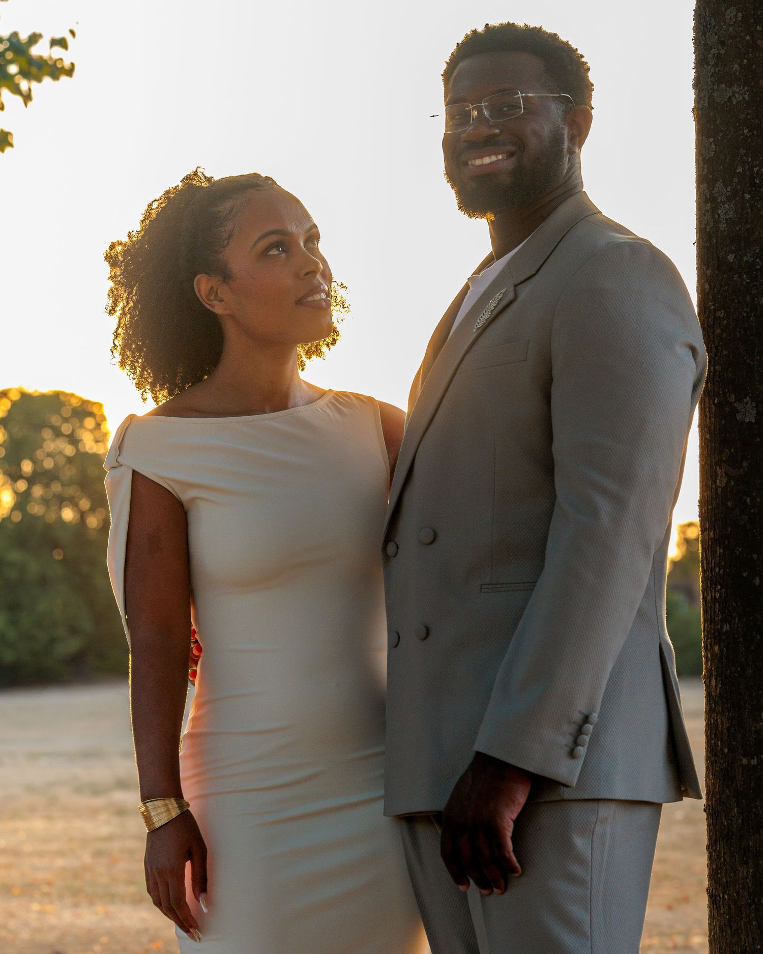 Elegant couple in wedding attire posing at golden hour outdoors, man in gray suit, woman in white dress.
