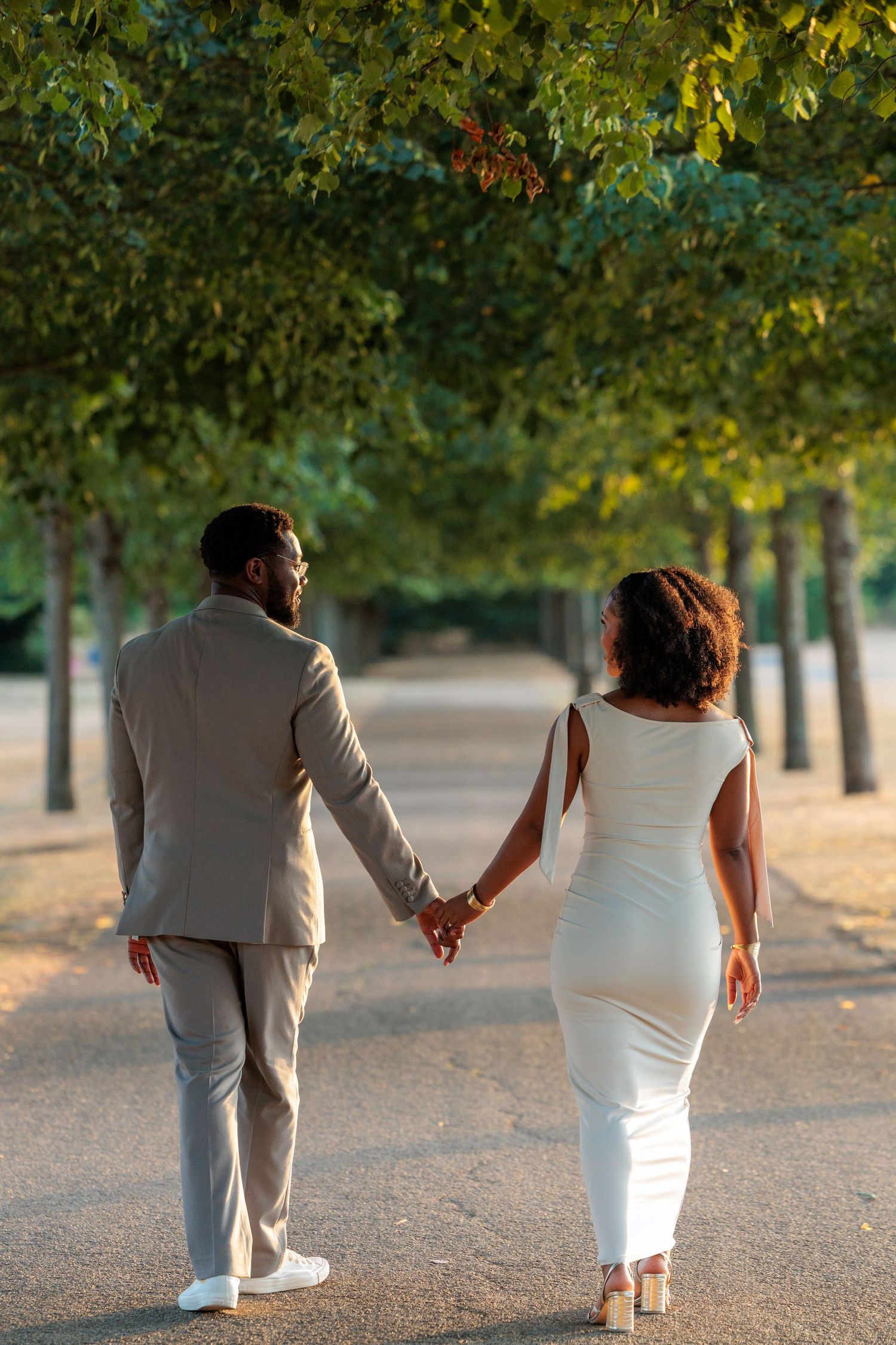 A couple holding hands walking down a tree-lined path, man in beige suit and woman in white dress.