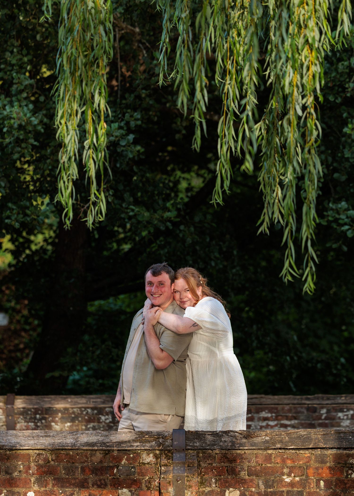 Couple embracing under weeping willow branches on a brick wall in a lush garden setting.