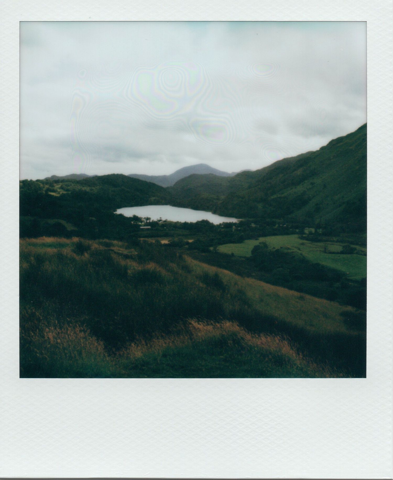 Moody Polaroid of a lush green valley with a lake nestled among rolling hills under overcast skies.