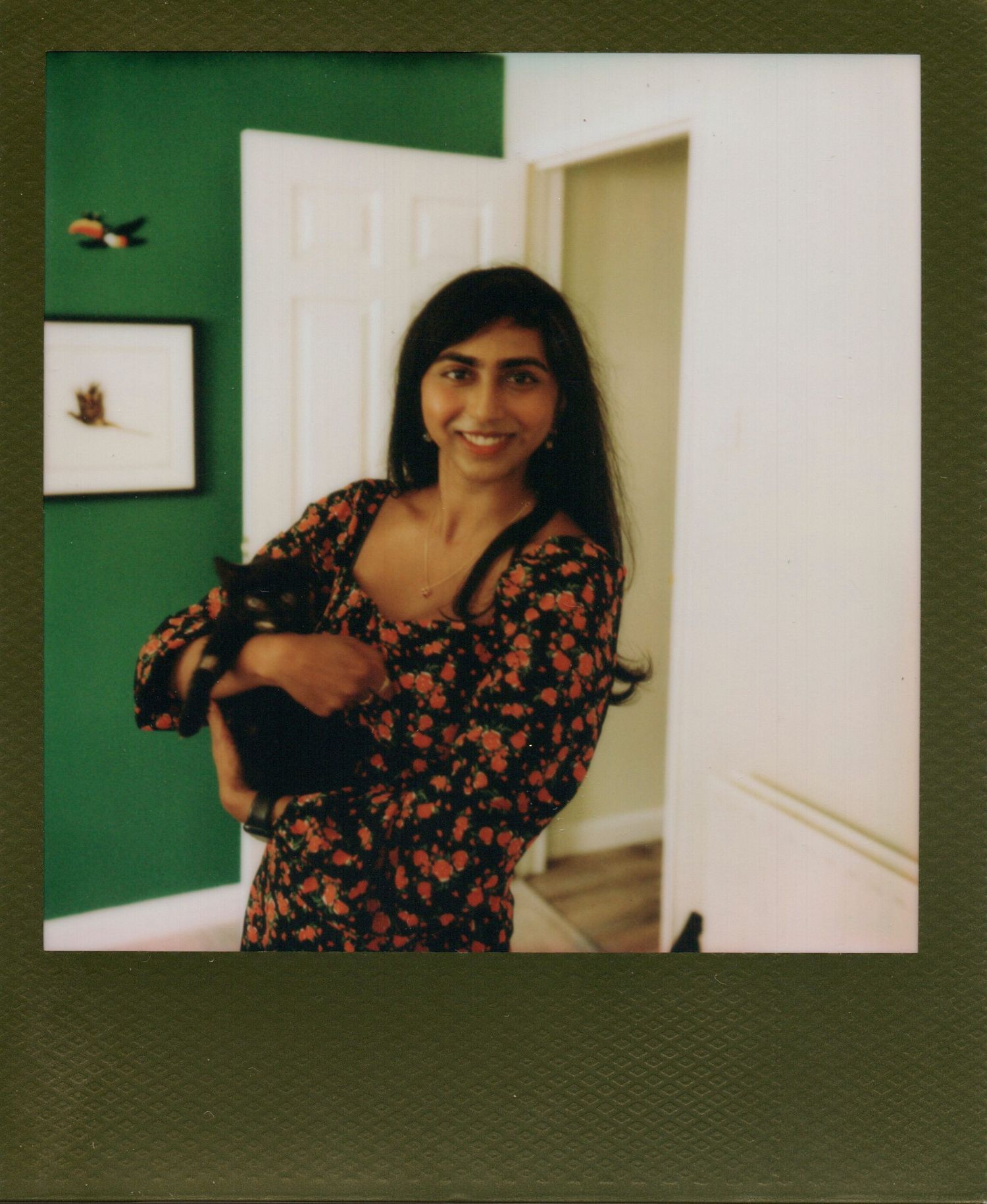 Smiling woman in floral dress holding a black cat in a green-walled room, Polaroid-style photo.