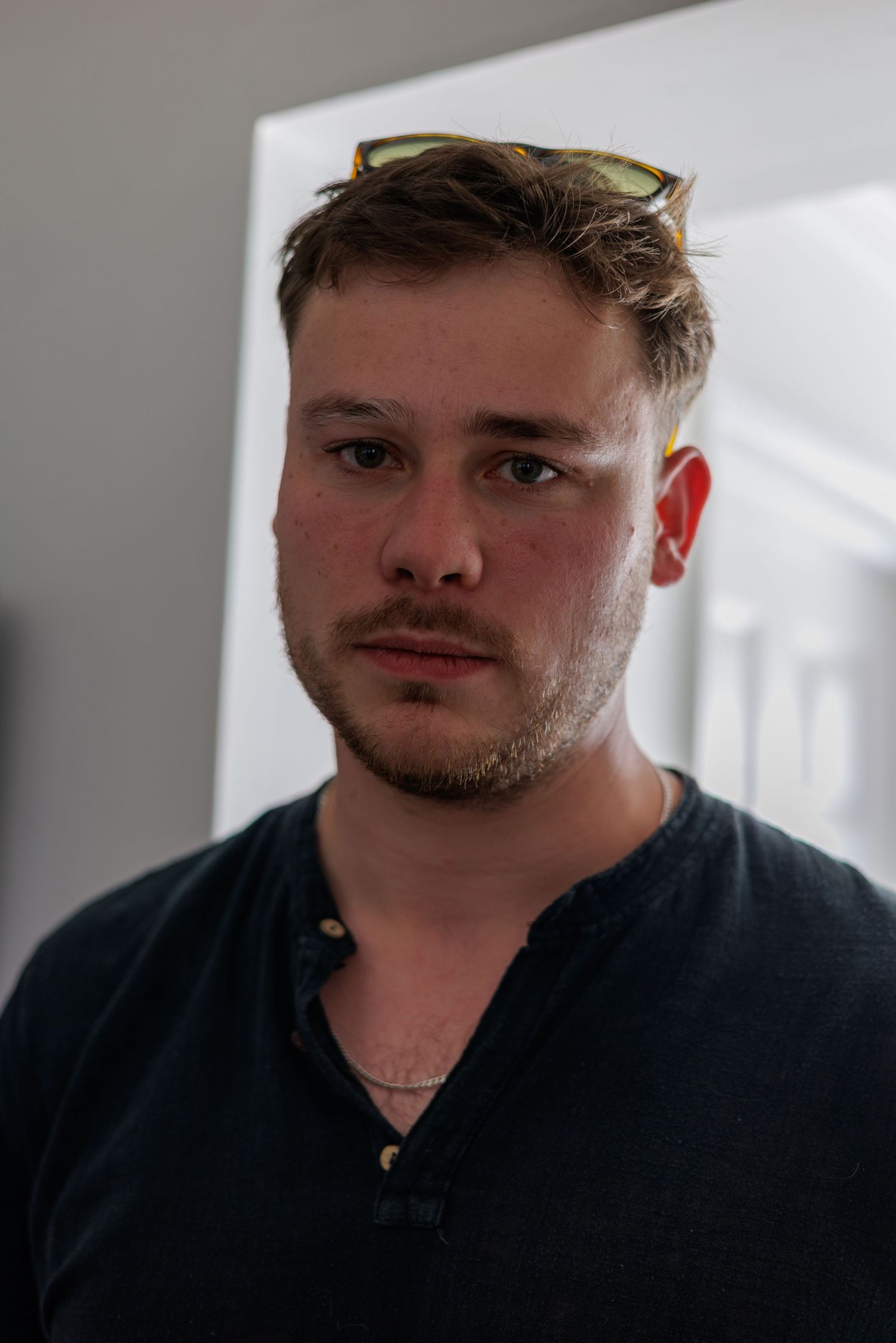 Young man with stubble and sunglasses on head wearing dark shirt, serious expression, indoor portrait photo.