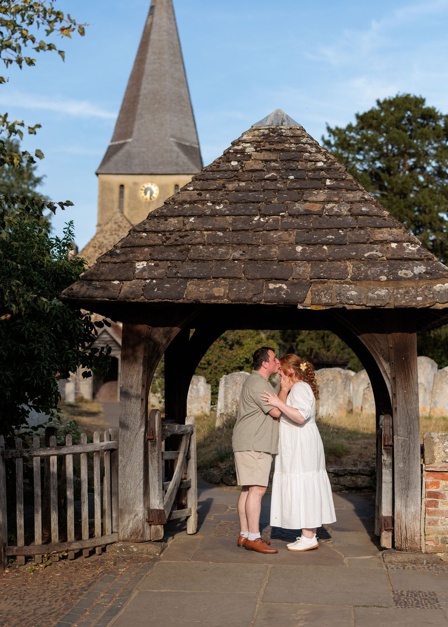 Couple sharing a kiss under a rustic lych-gate at a historic English church with a stone steeple.