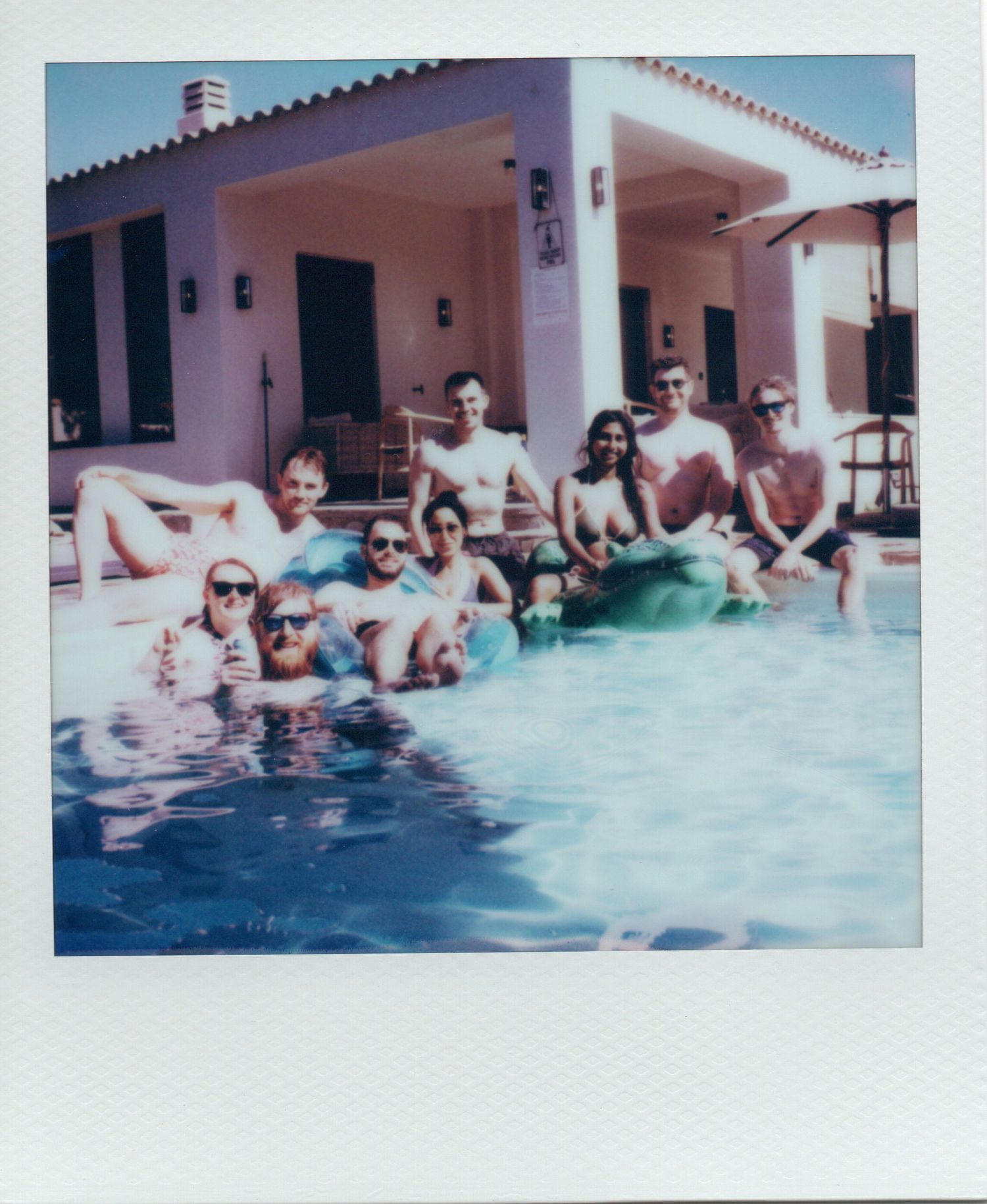 Group of friends relaxing in a sunny outdoor swimming pool beside a Mediterranean-style villa.
