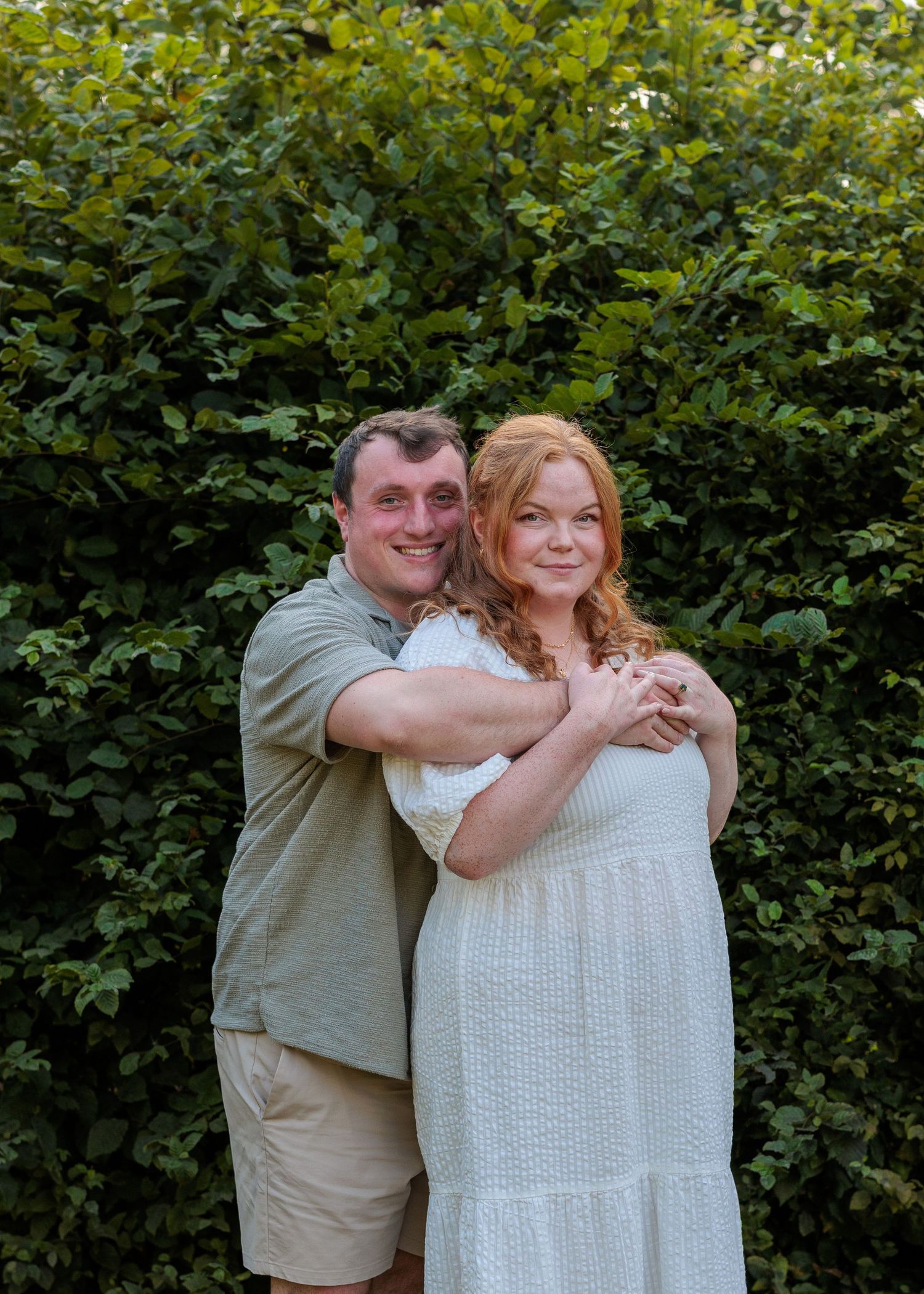 Couple embracing and smiling in front of lush green hedge, woman in white dress, man in khaki outfit.