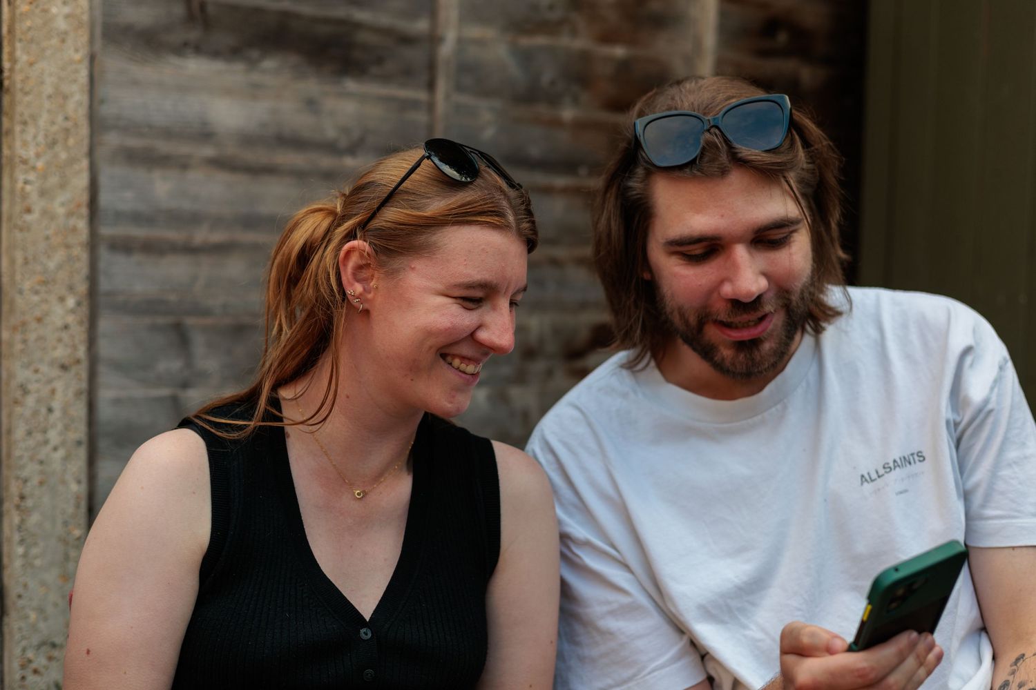 A smiling woman and bearded man with sunglasses look at a phone together outdoors.