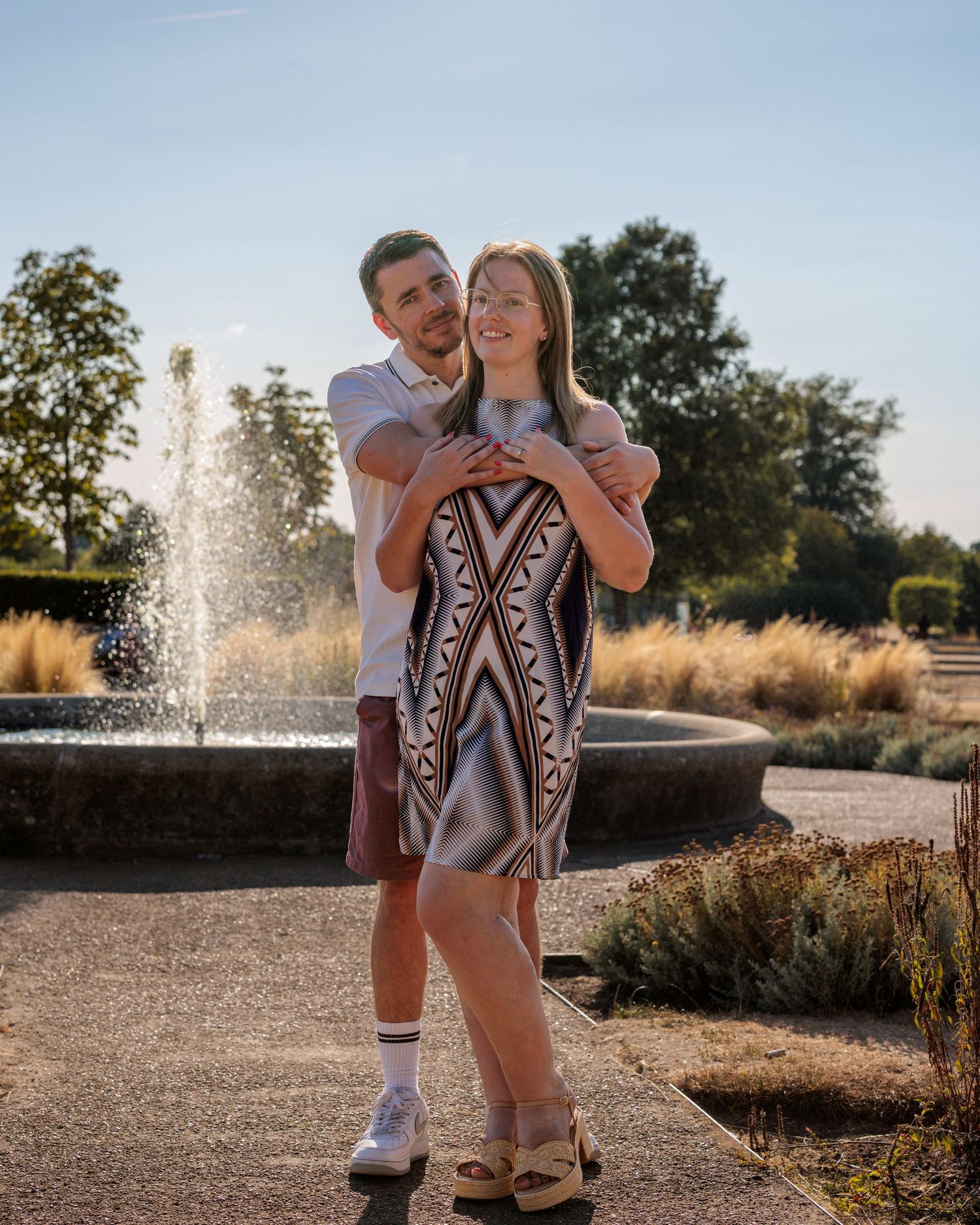 Couple embracing near a fountain in a sunny park, woman in patterned dress, man in white shirt and shorts.