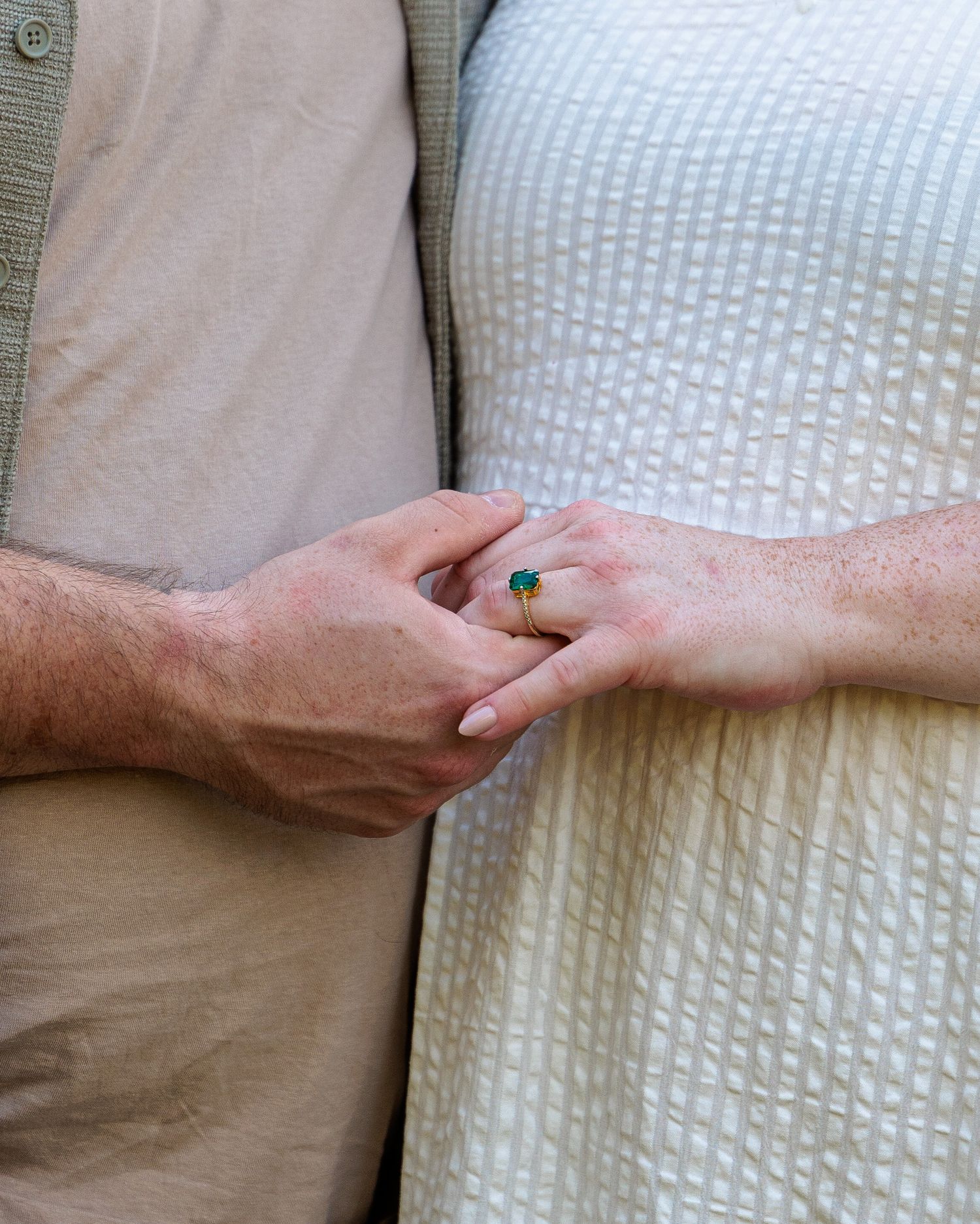 Close-up of couple holding hands, woman wearing gold ring with teal gemstone, man in khaki shirt, woman in white dress.