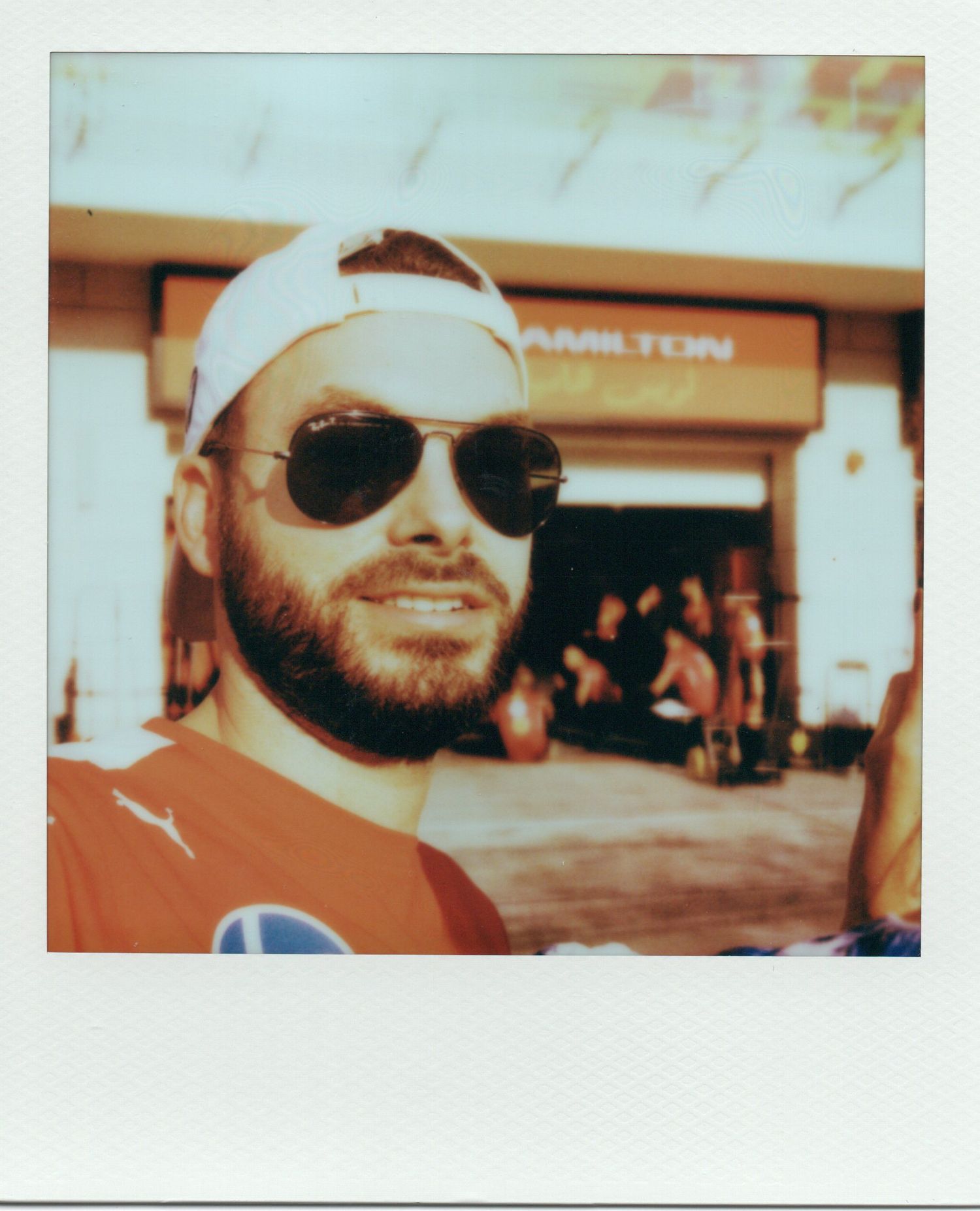 Bearded man in sunglasses and cap smiling at a Formula 1 Hamilton pit lane event.