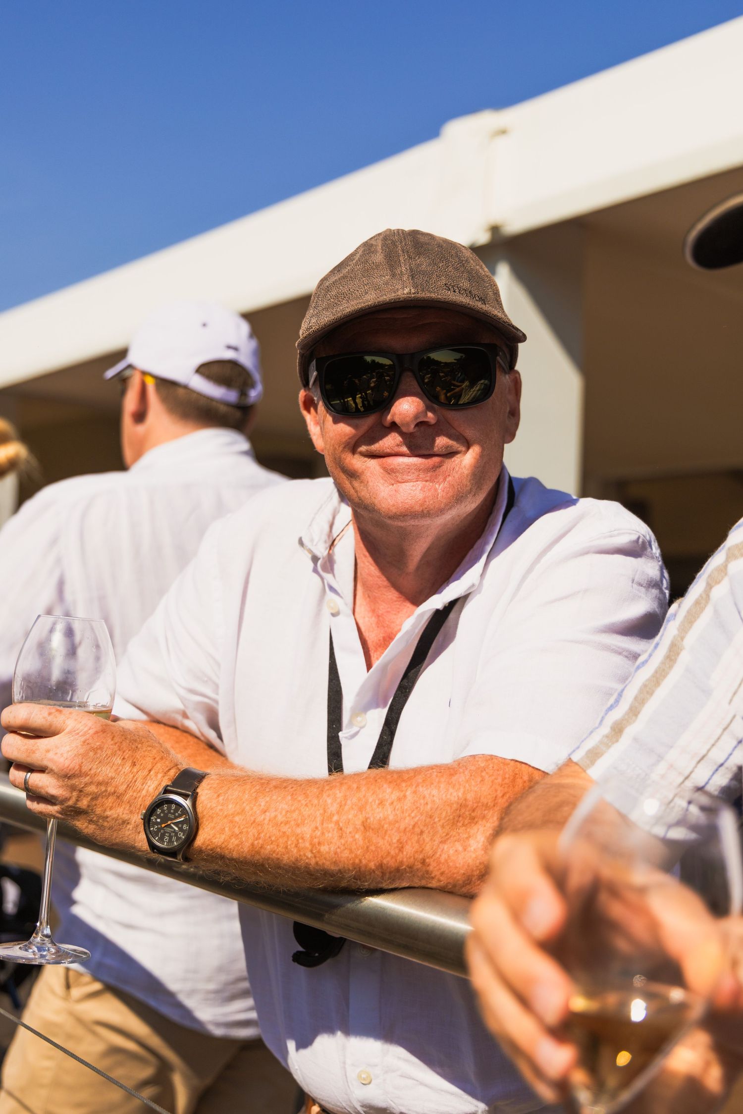 Man in cap and sunglasses smiling while leaning on railing with wine glass at outdoor event.