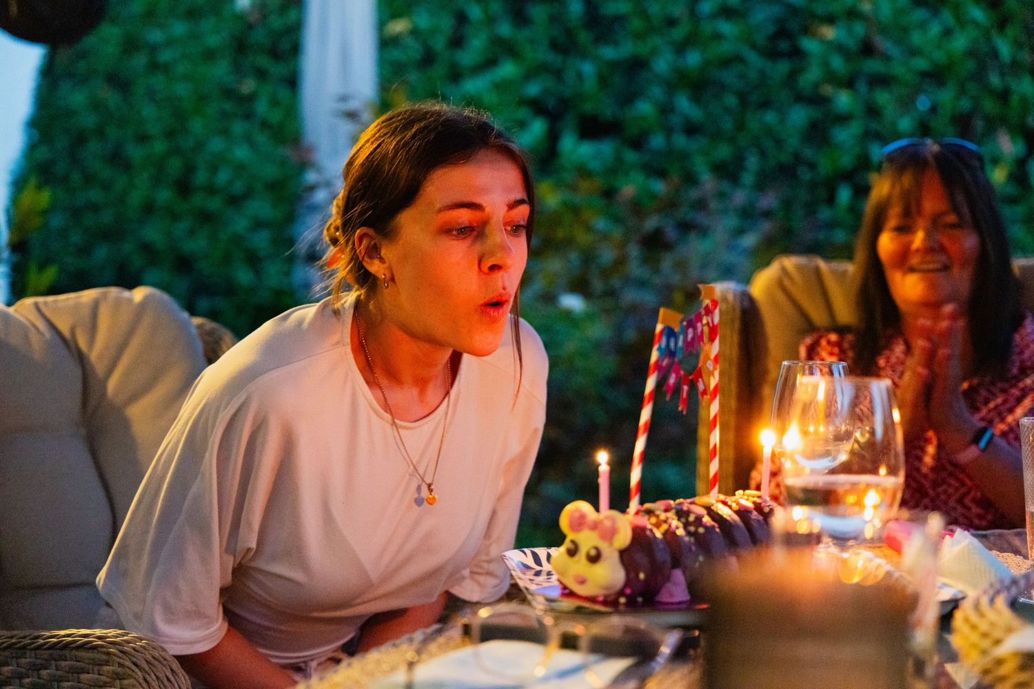 Young woman blowing out birthday cake candles at an outdoor evening celebration with guests nearby.