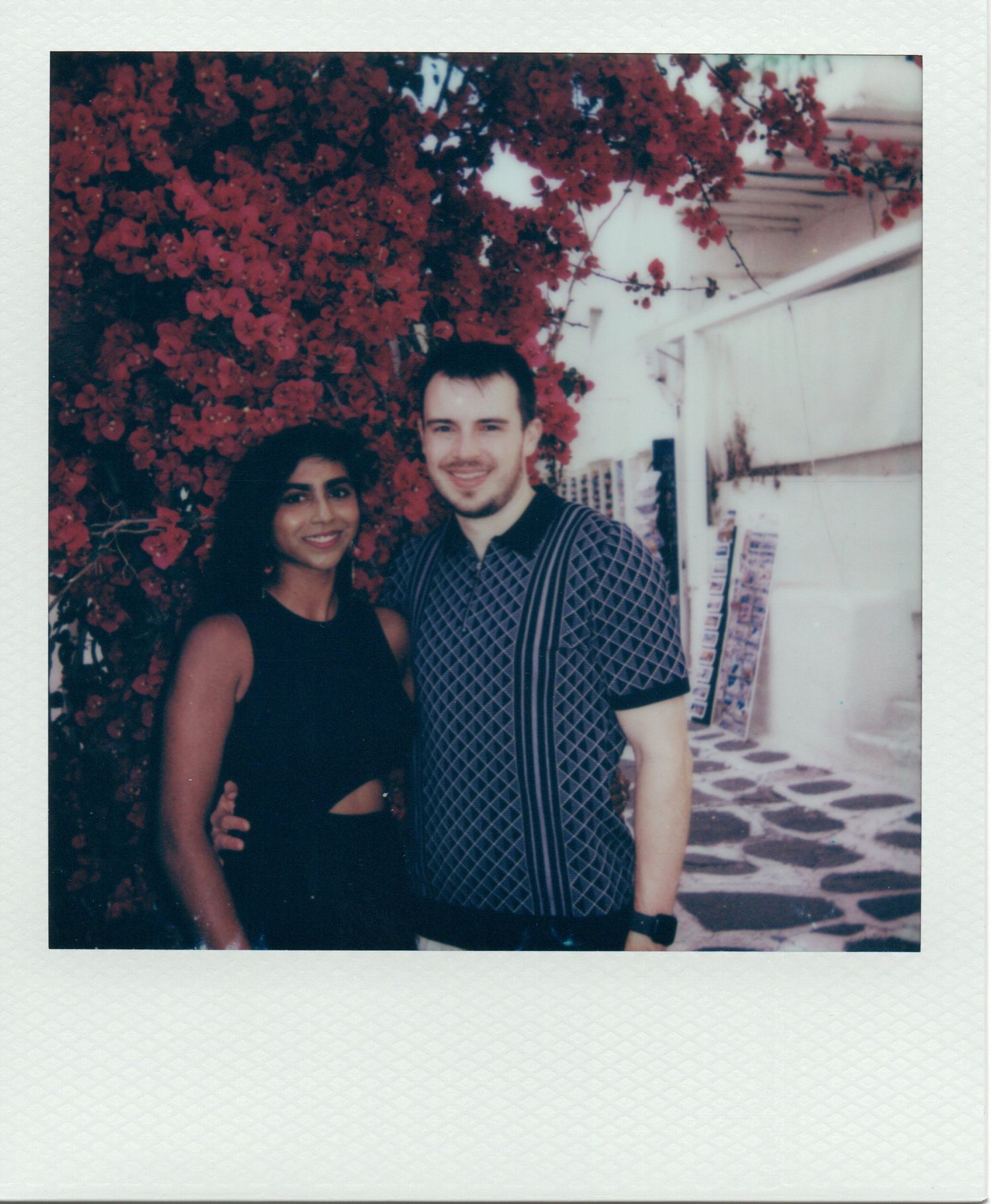 Couple smiling together in front of vibrant red bougainvillea on a sunny street.