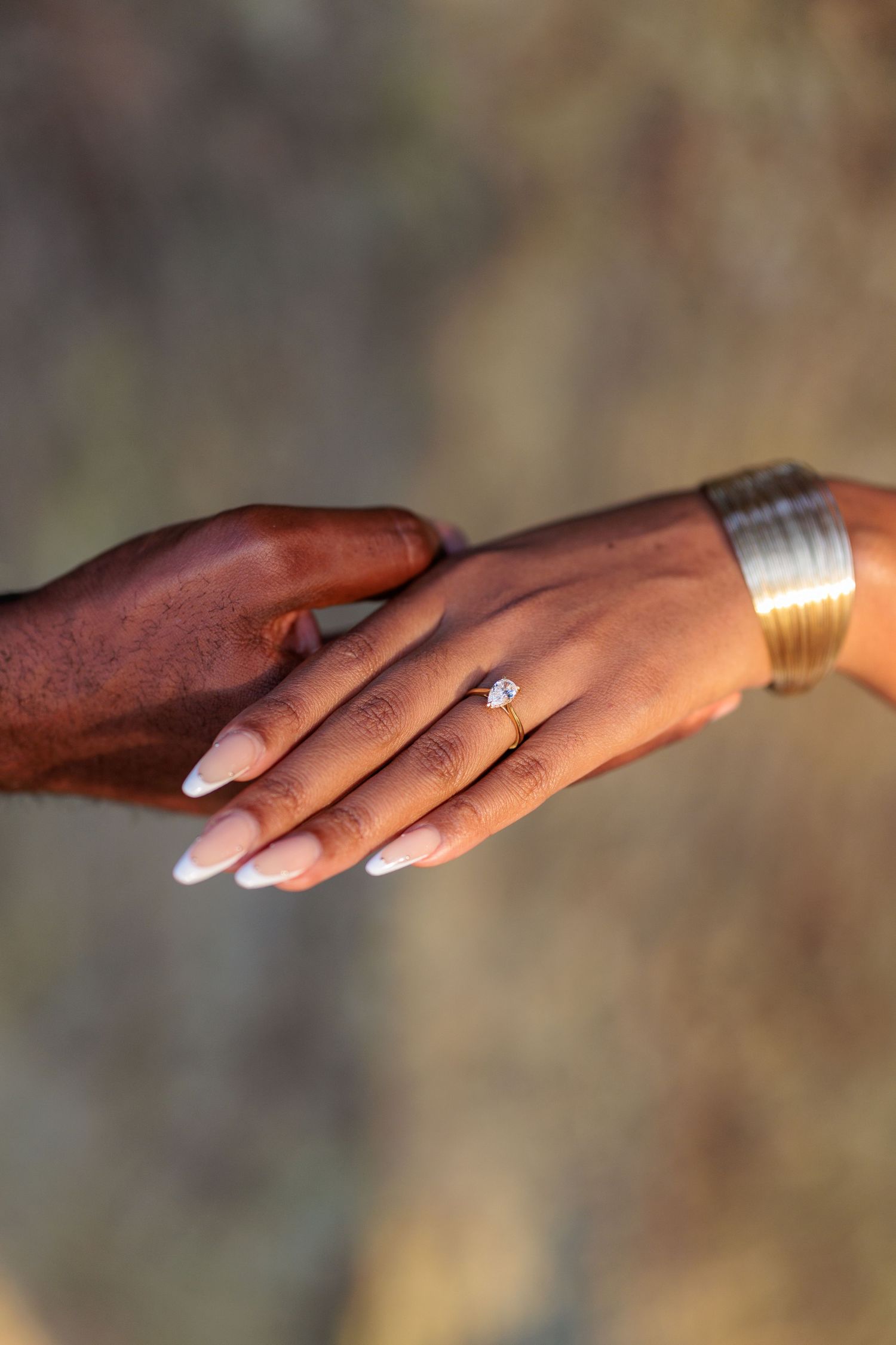 Two hands holding, one with a diamond engagement ring and gold bracelet, symbolizing love and commitment.
