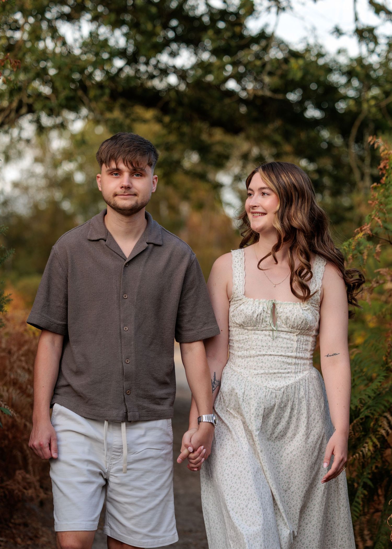 A young couple holds hands while walking on a nature path, the woman in a floral dress and man in a gray shirt and white shorts.