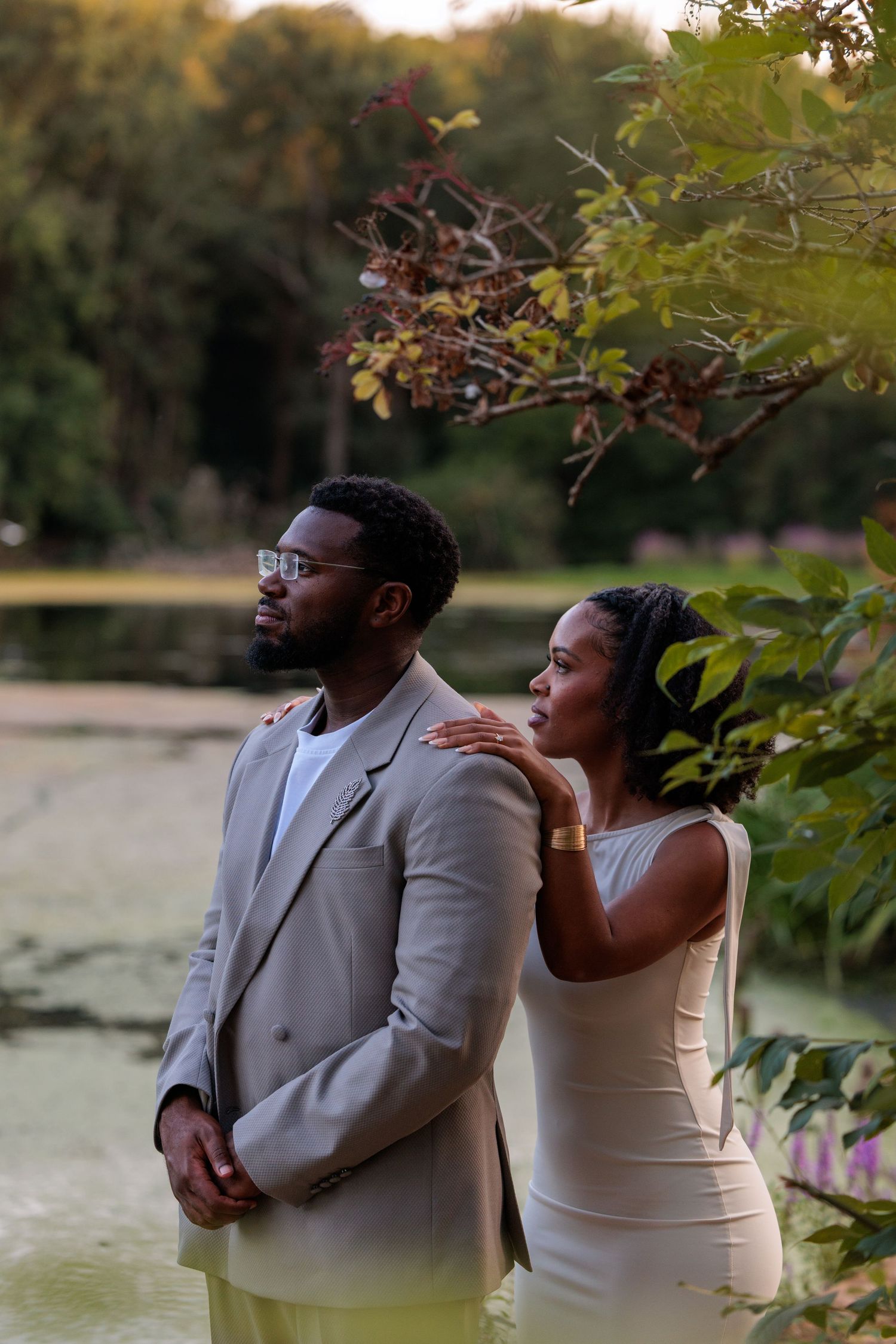 A couple poses by a serene pond, man in gray suit, woman in cream dress resting hand on his shoulder.