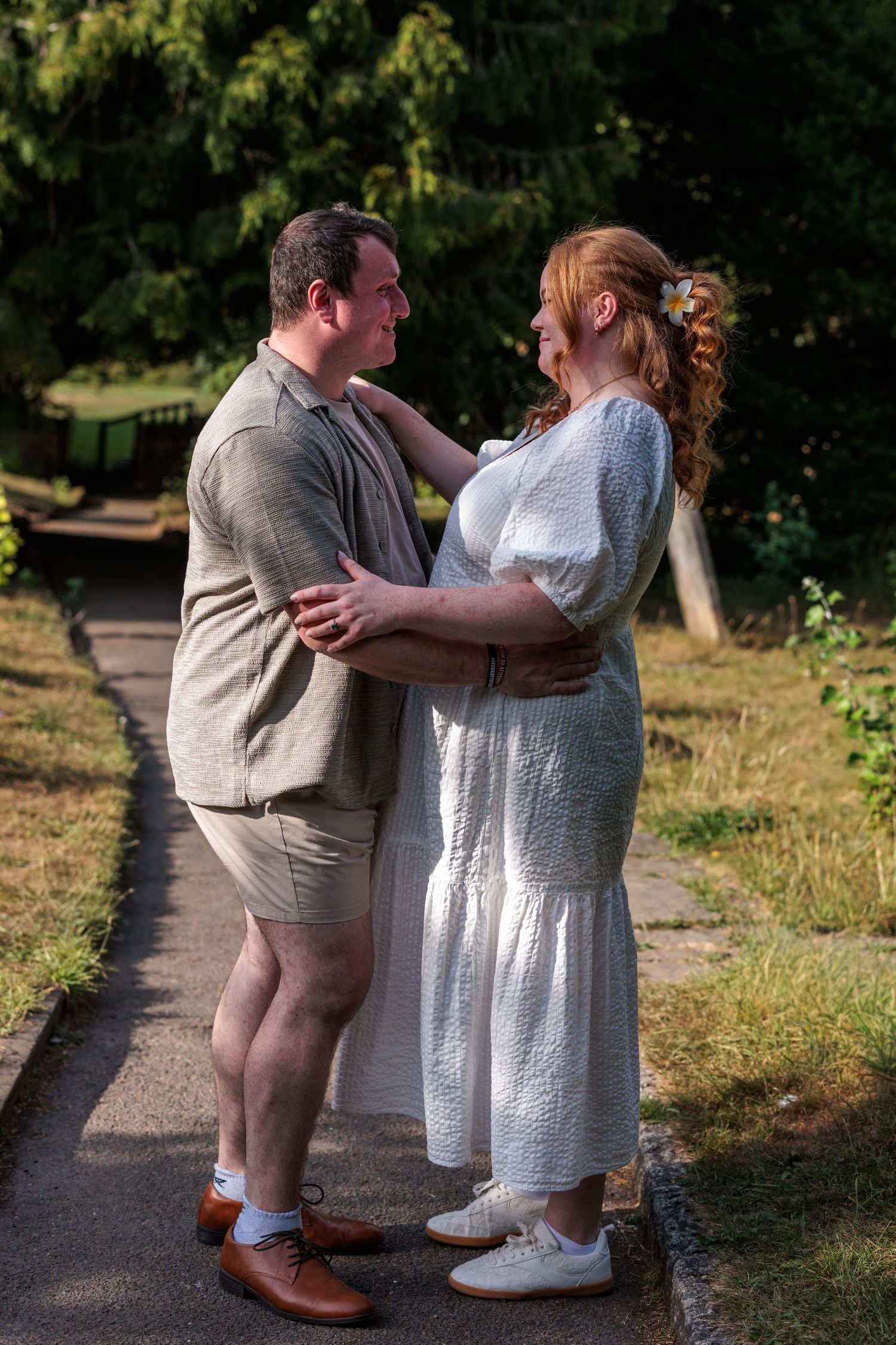 Couple embracing outdoors on a sunny path, woman in white dress and man in linen shorts, surrounded by green trees.