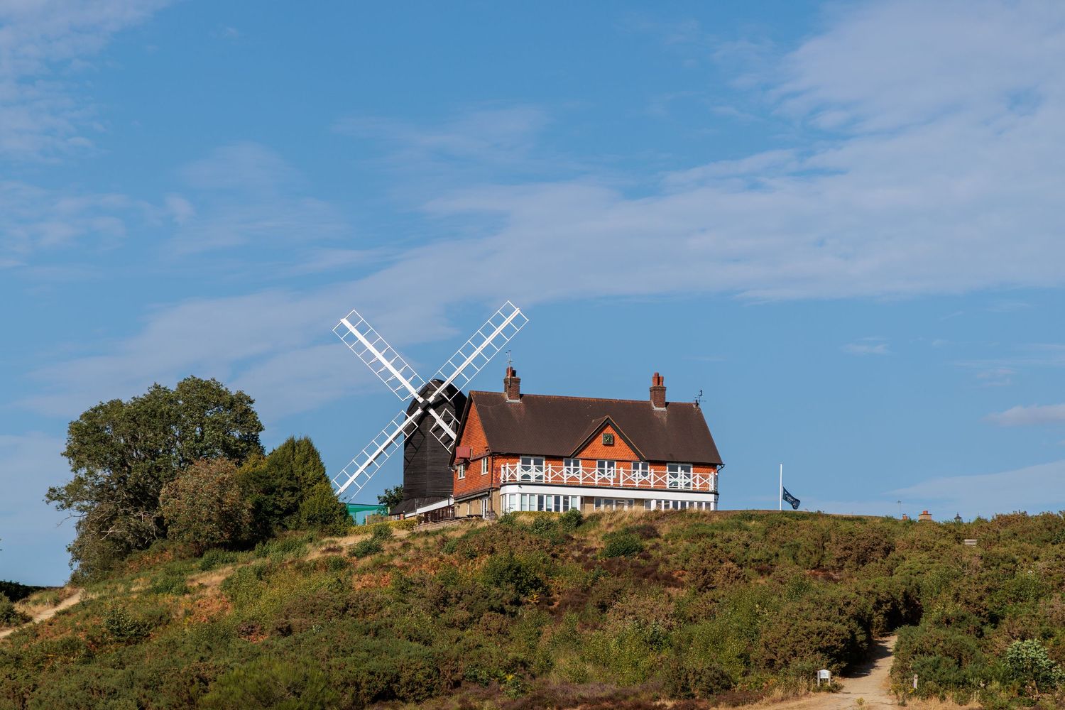 A traditional windmill beside a red and white timber-framed house on a hill under a blue sky.
