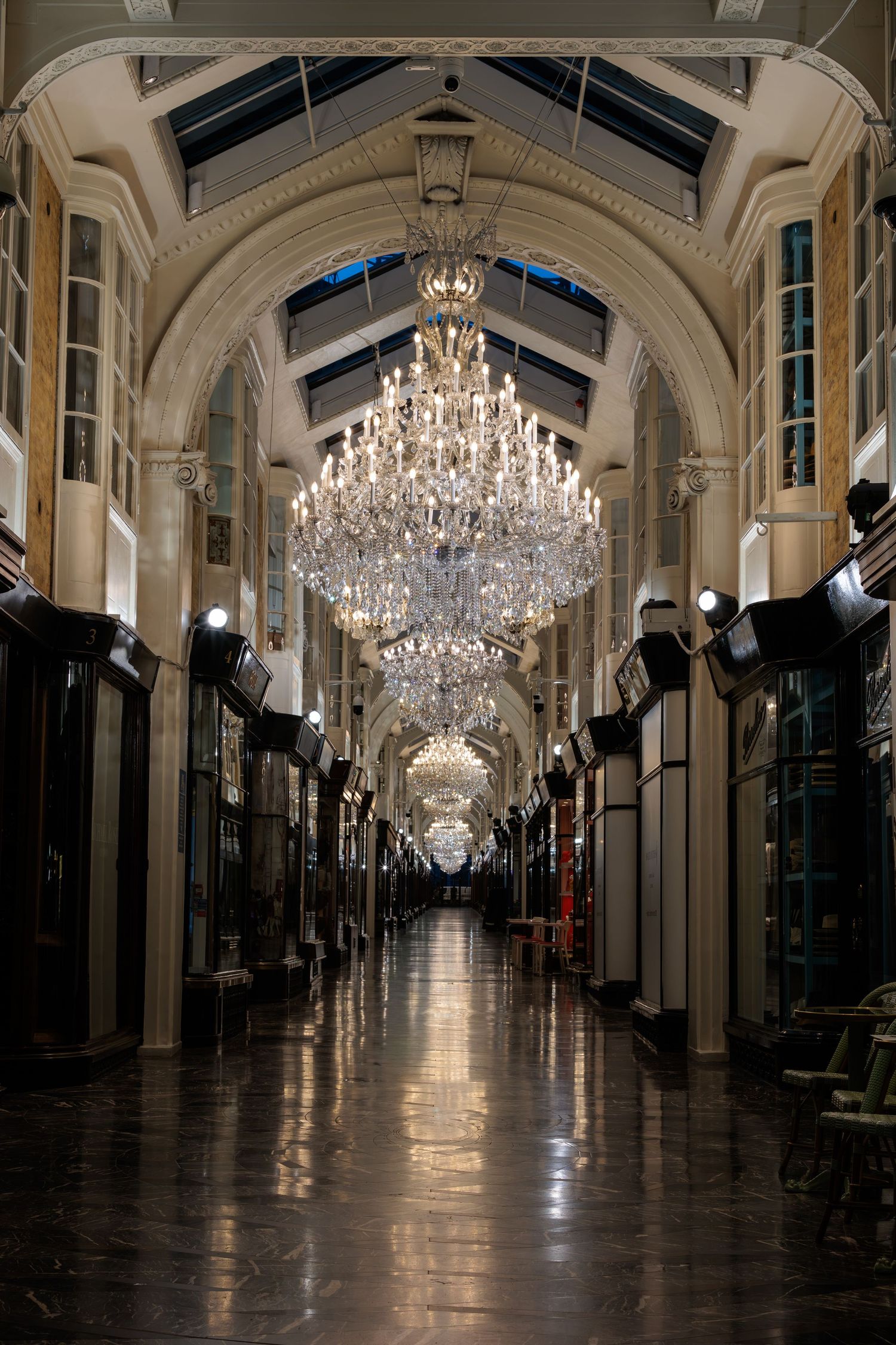 Elegant indoor shopping arcade with crystal chandeliers, arched ceilings, and reflective marble floors in a grand Victorian-style passage.