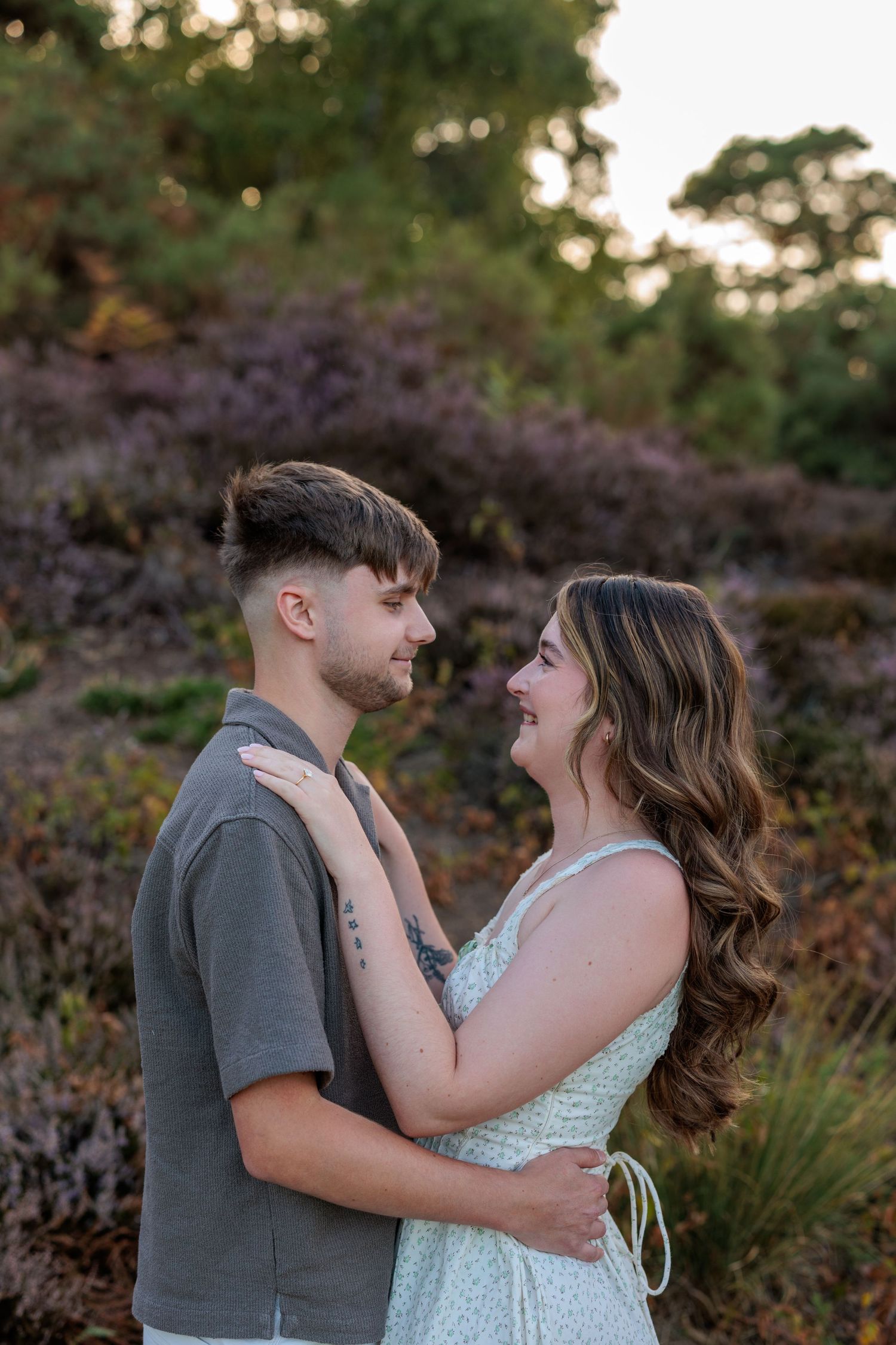 A couple embraces and gazes at each other in a scenic outdoor setting with purple heather and green trees at sunset.