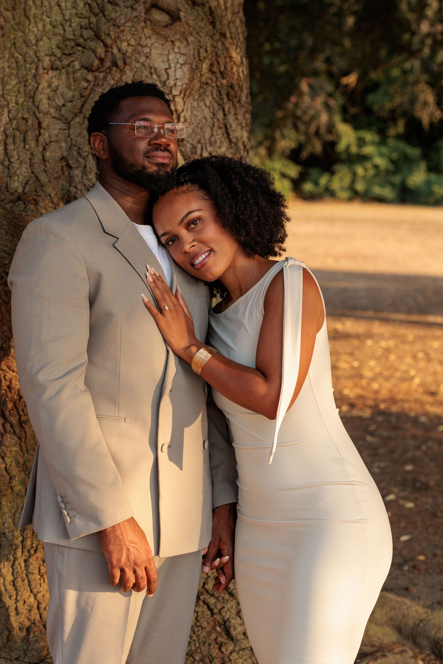Elegant Black couple in matching white and beige outfits pose romantically by a tree in a sunlit park.