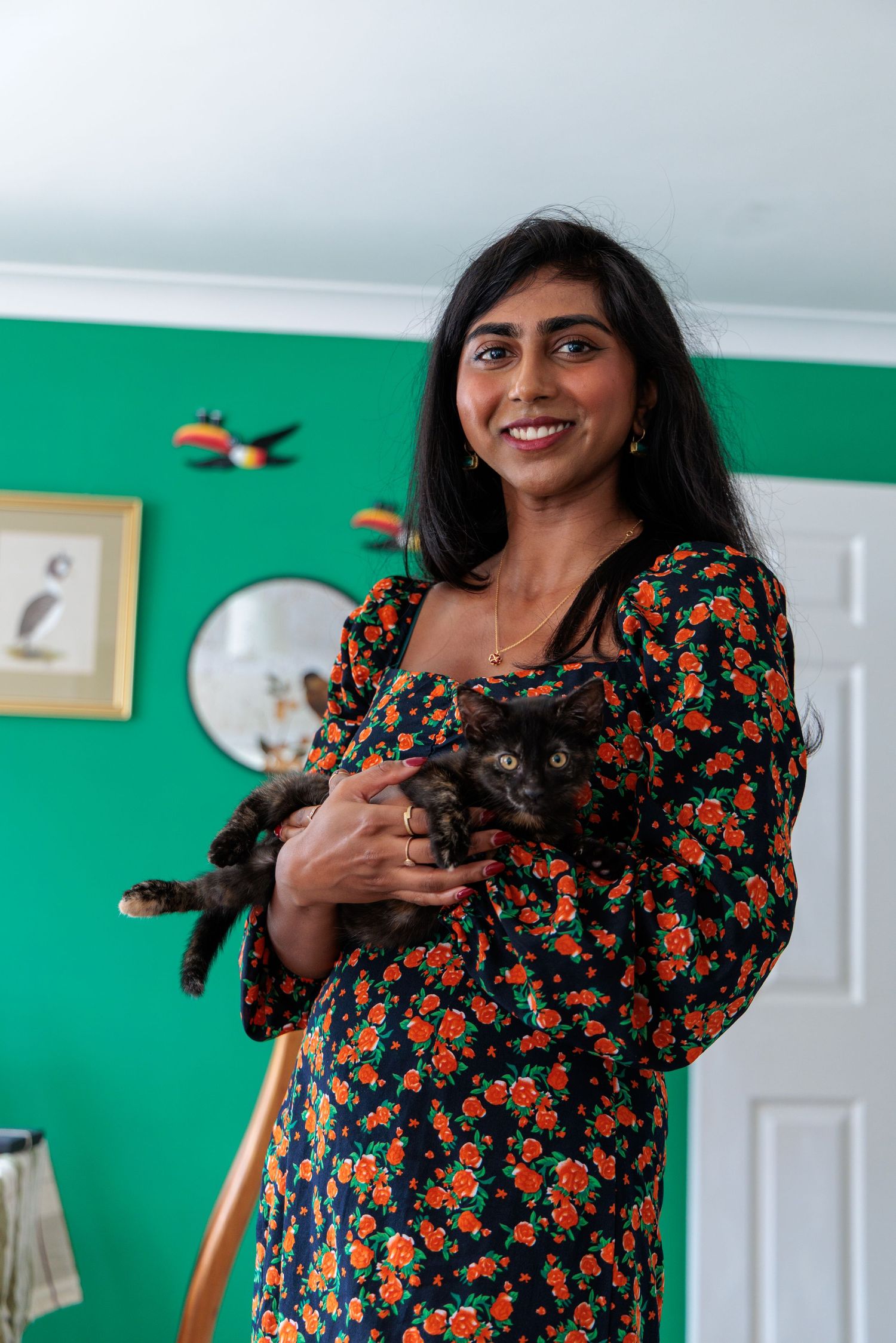 Smiling woman in floral dress holding a dark tortoiseshell cat in a green-walled room with bird wall art.