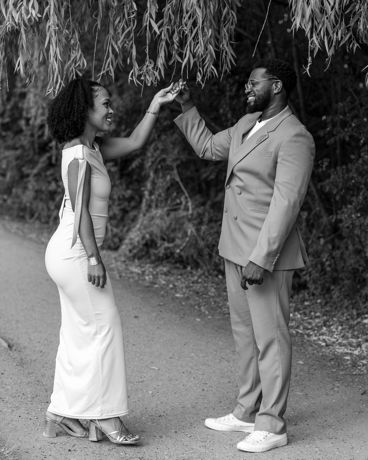 Black and white photo of a couple dancing outdoors, woman in white dress, man in suit, smiling joyfully.