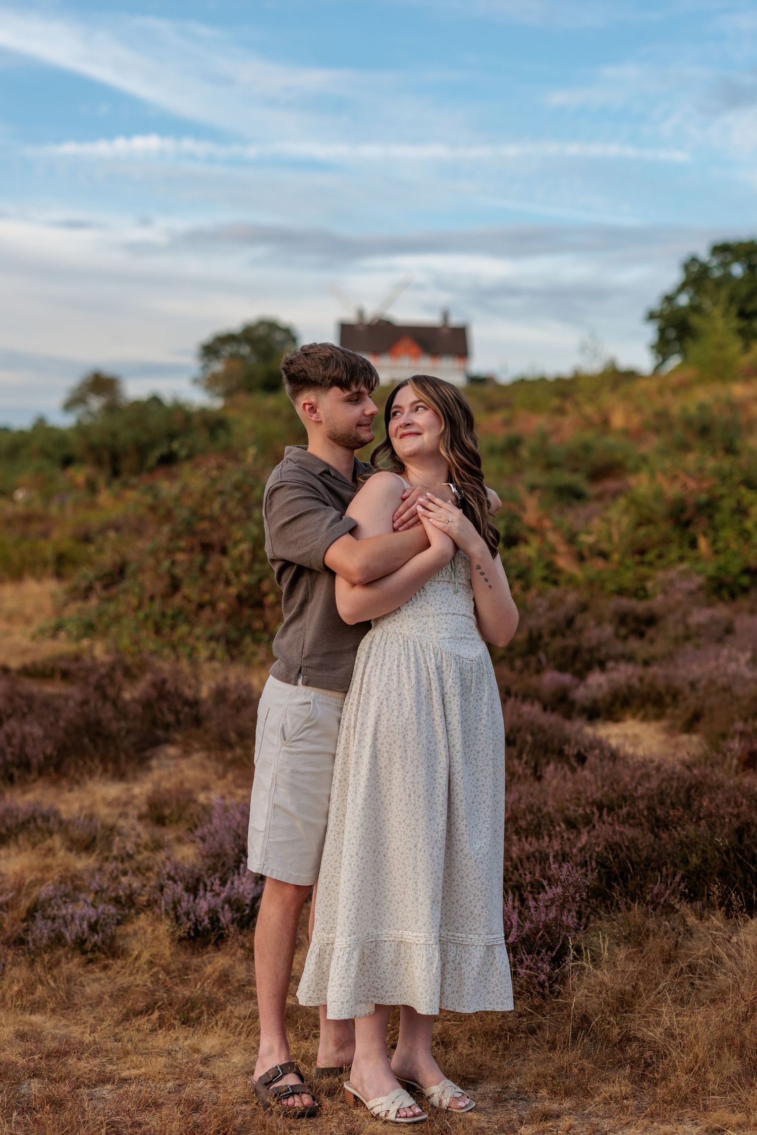 A couple embraces in a heather field at golden hour, the woman in a white dress smiling up at the man holding her.