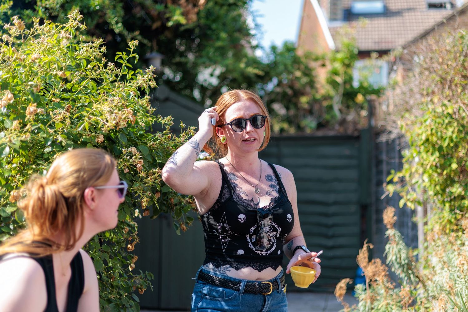 Two women outdoors on a sunny day, one in a black tank top with sunglasses holding fruit in a garden alley.