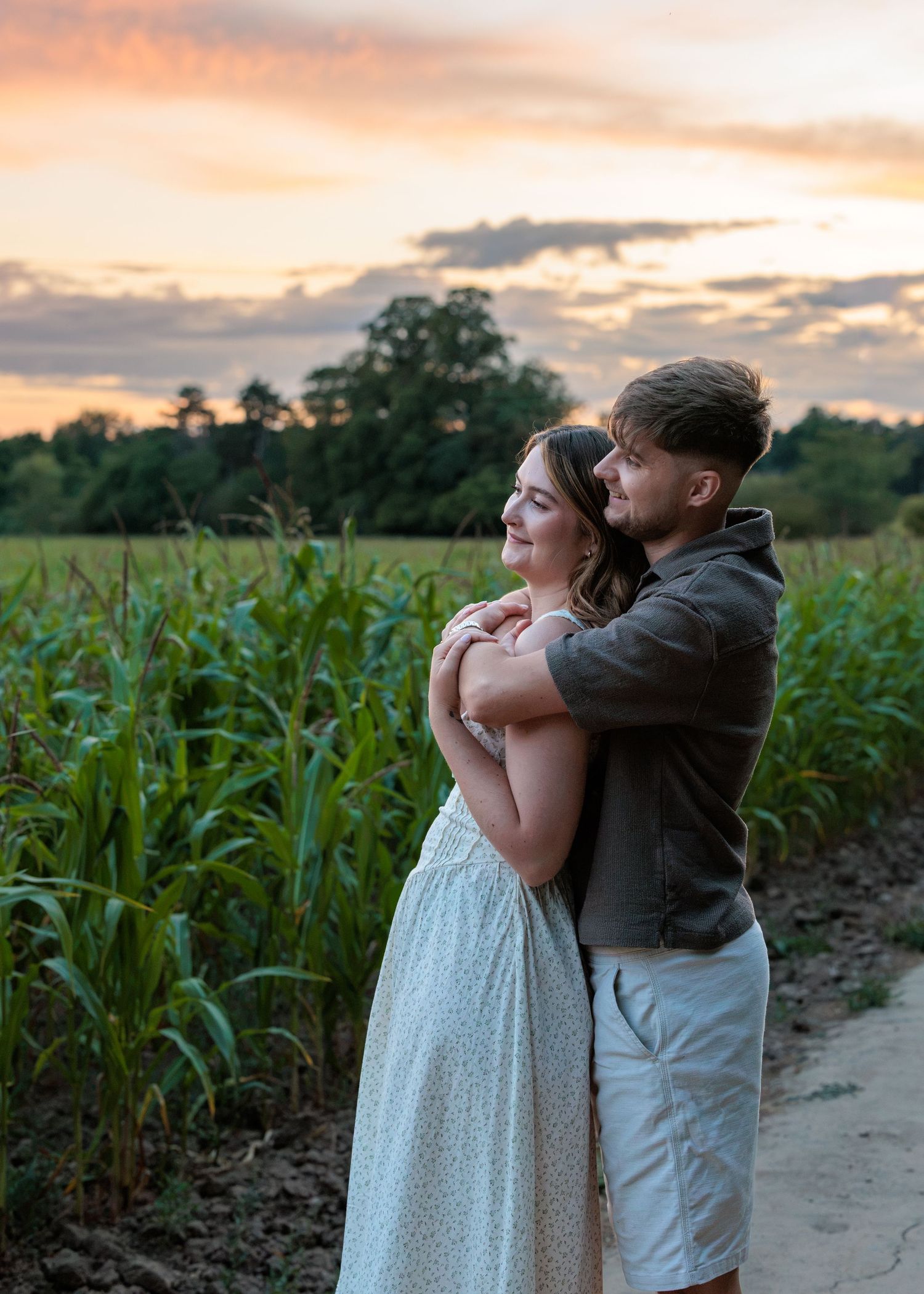 Couple embracing at sunset near a cornfield, woman in white dress, man in gray jacket, colorful sky backdrop.