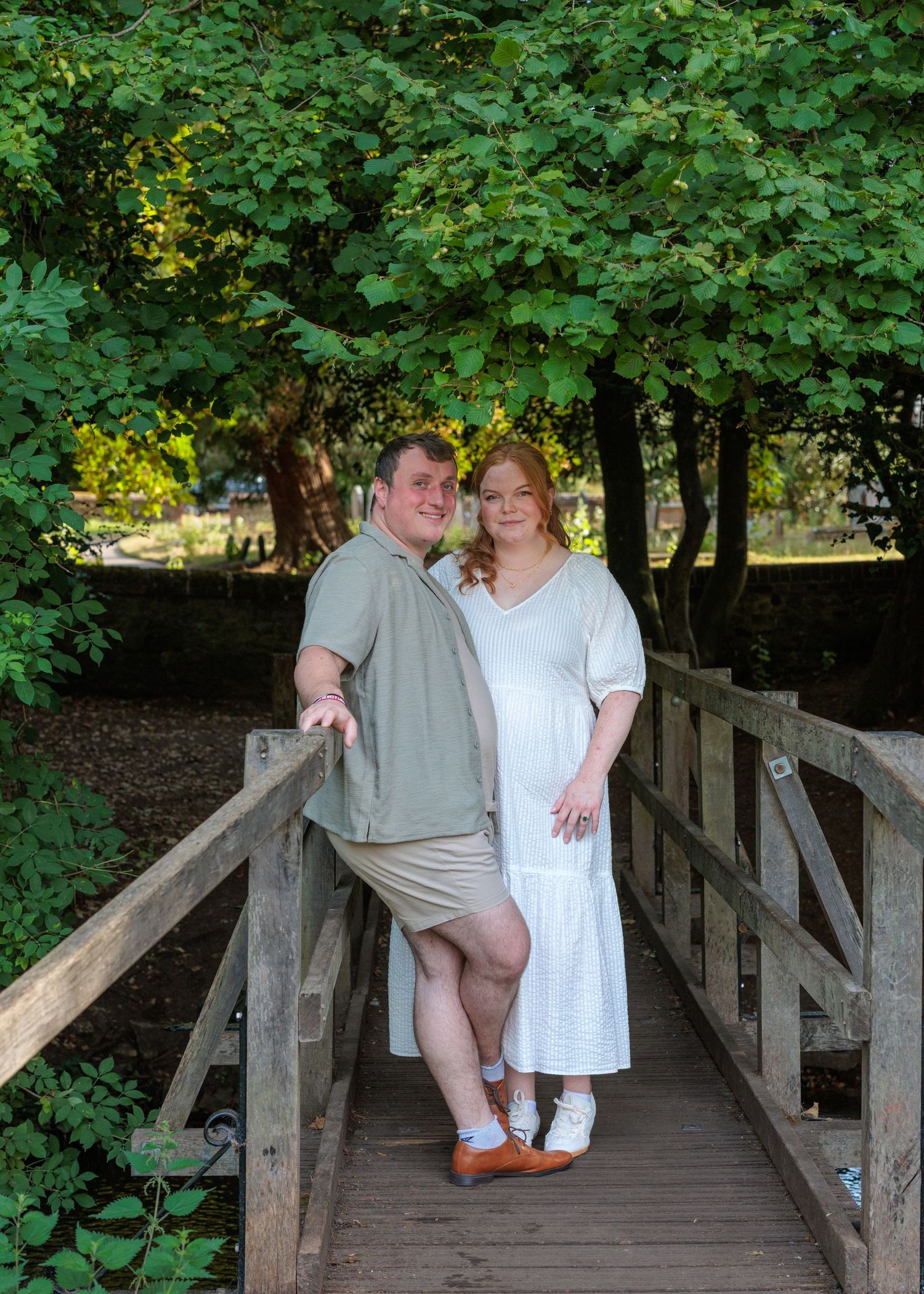 Couple posing on a wooden bridge surrounded by lush green trees in an outdoor park setting.