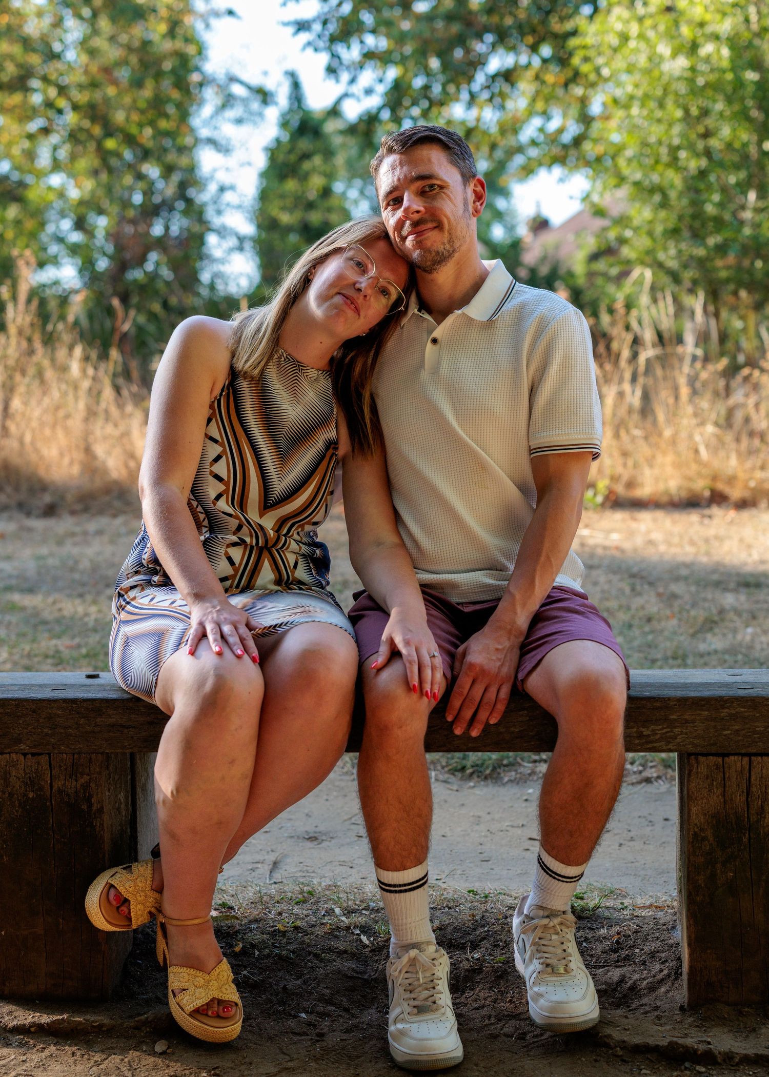 A couple sits closely on a wooden bench outdoors, smiling warmly, surrounded by trees in a sunny park setting.