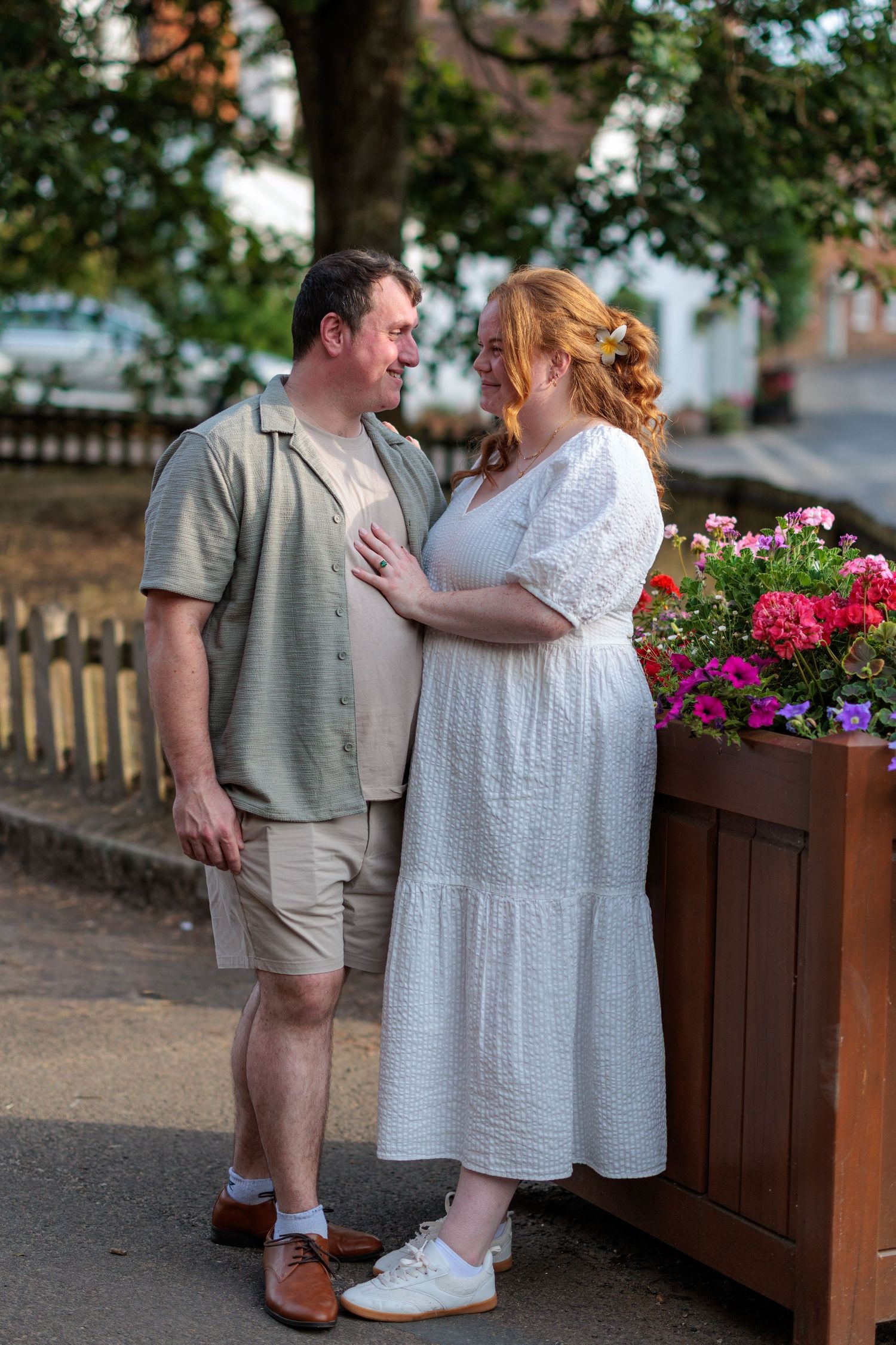A couple in casual attire stands together near a flower planter outdoors, sharing a warm smile on a summer day.