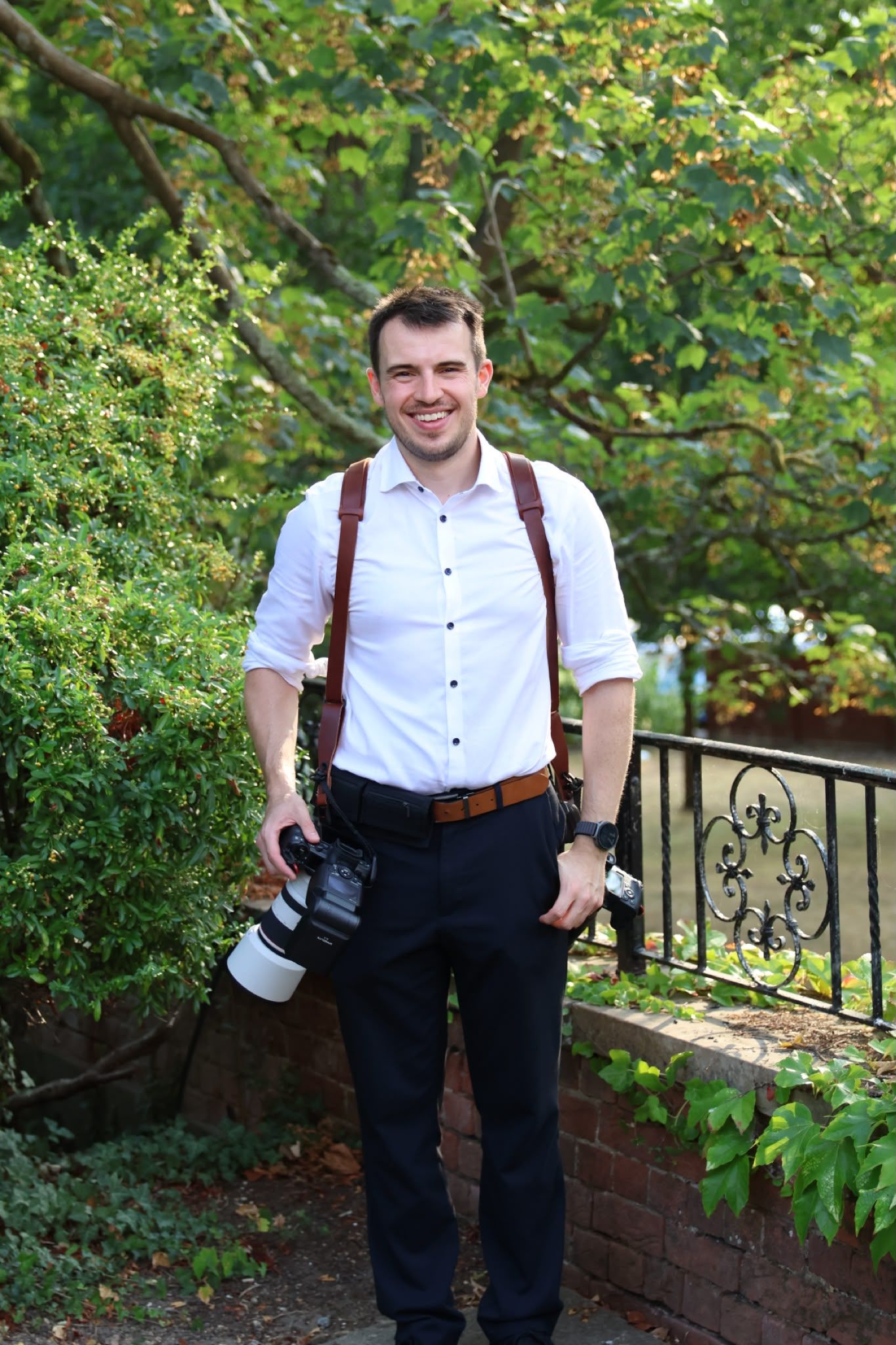 Smiling male photographer in white shirt and suspenders holding camera outdoors near green trees and iron fence.