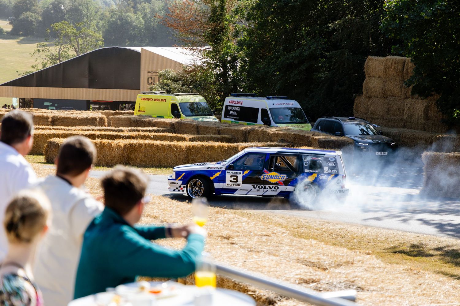 Blue rally car drifting with smoke at an outdoor motorsport event, with hay bales lining the track and spectators watching.