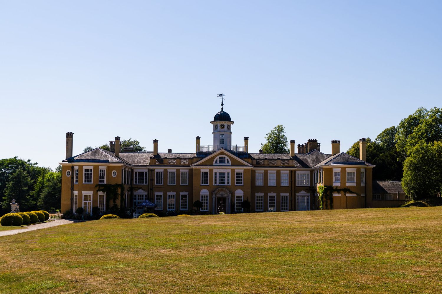 Grand Georgian country house with clock tower cupola set in expansive dry summer lawns under clear blue sky.