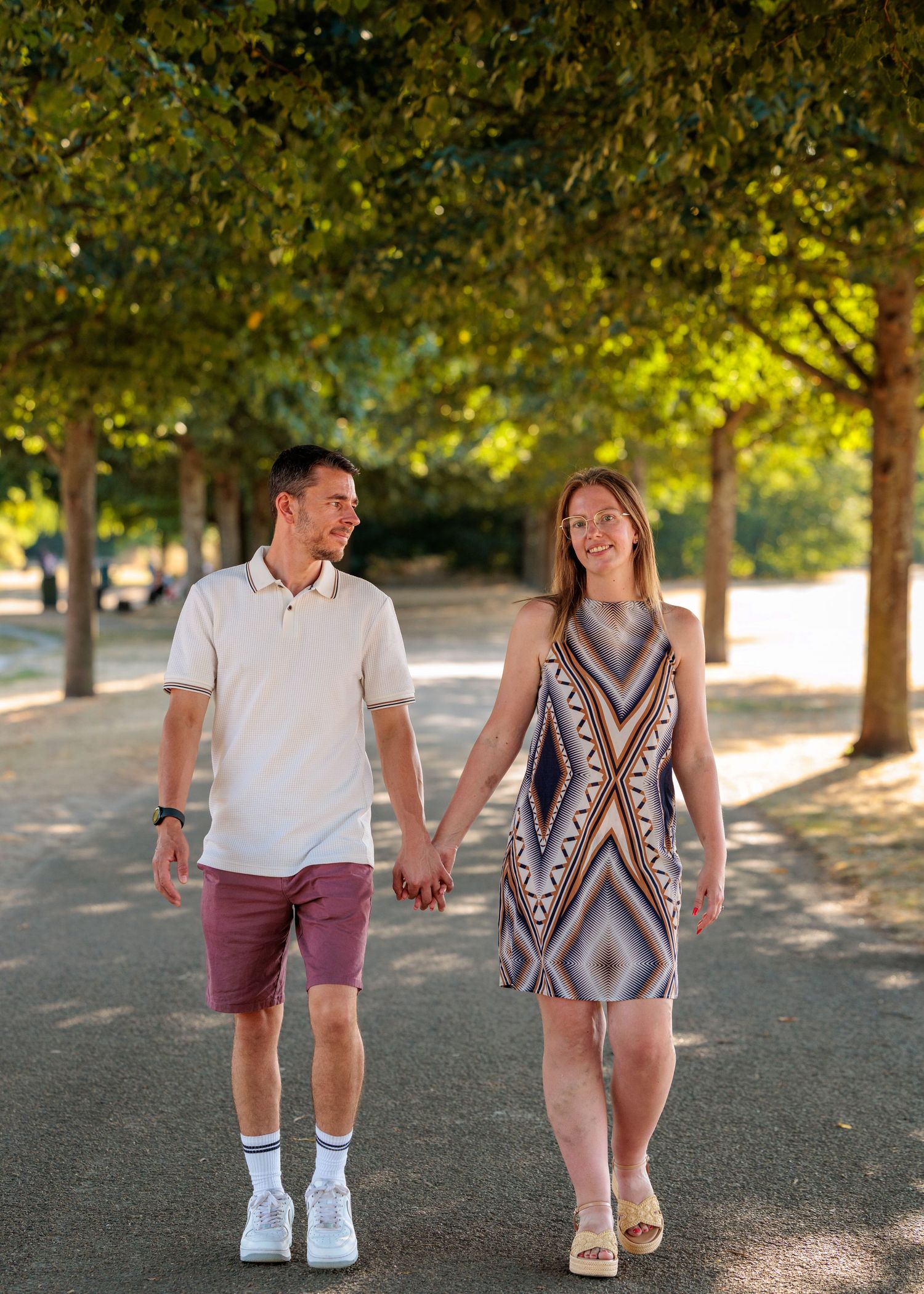 A smiling couple holds hands while walking through a tree-lined park path on a sunny day.