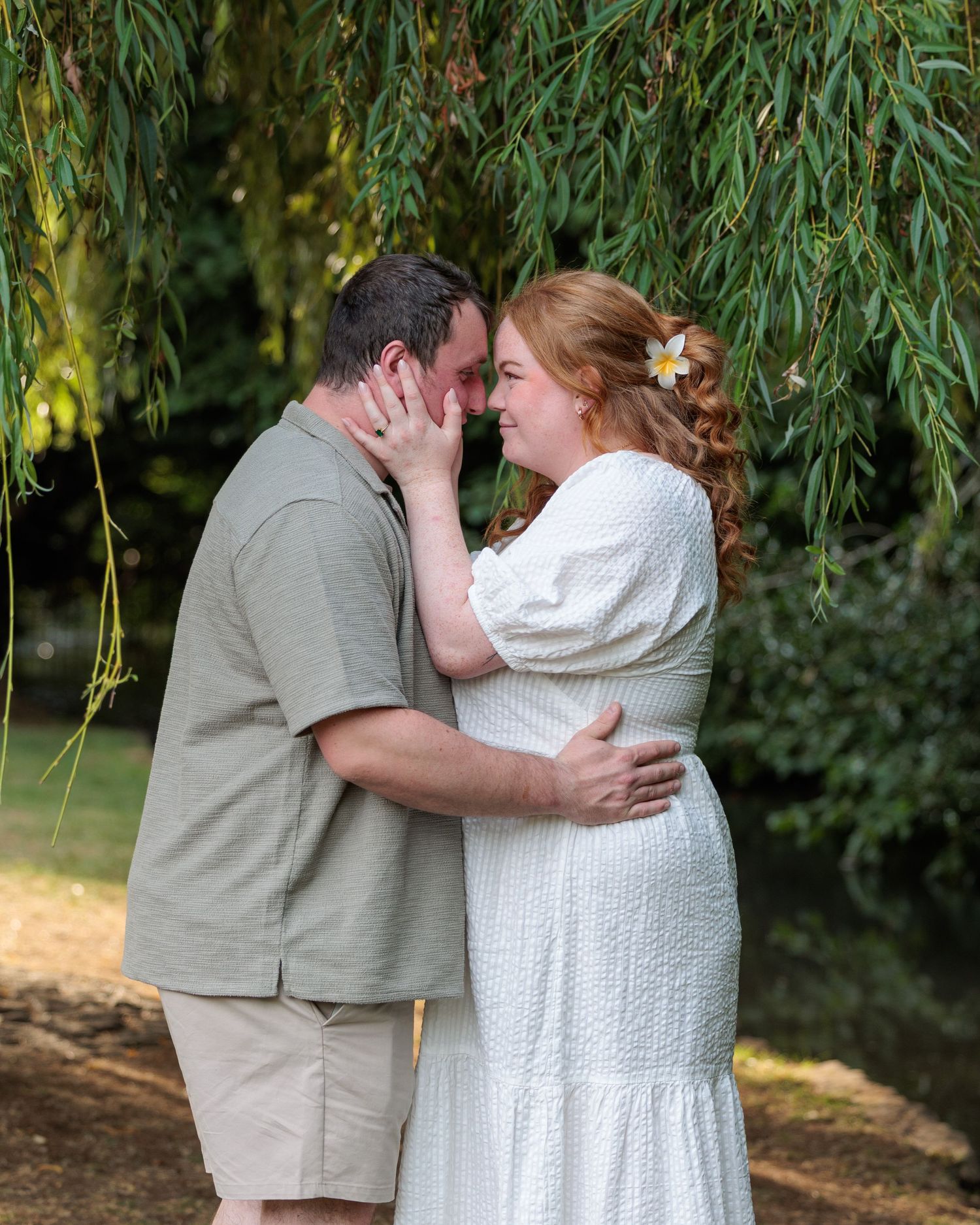 Couple sharing a romantic moment under a weeping willow tree, bride in white dress with flower in hair.