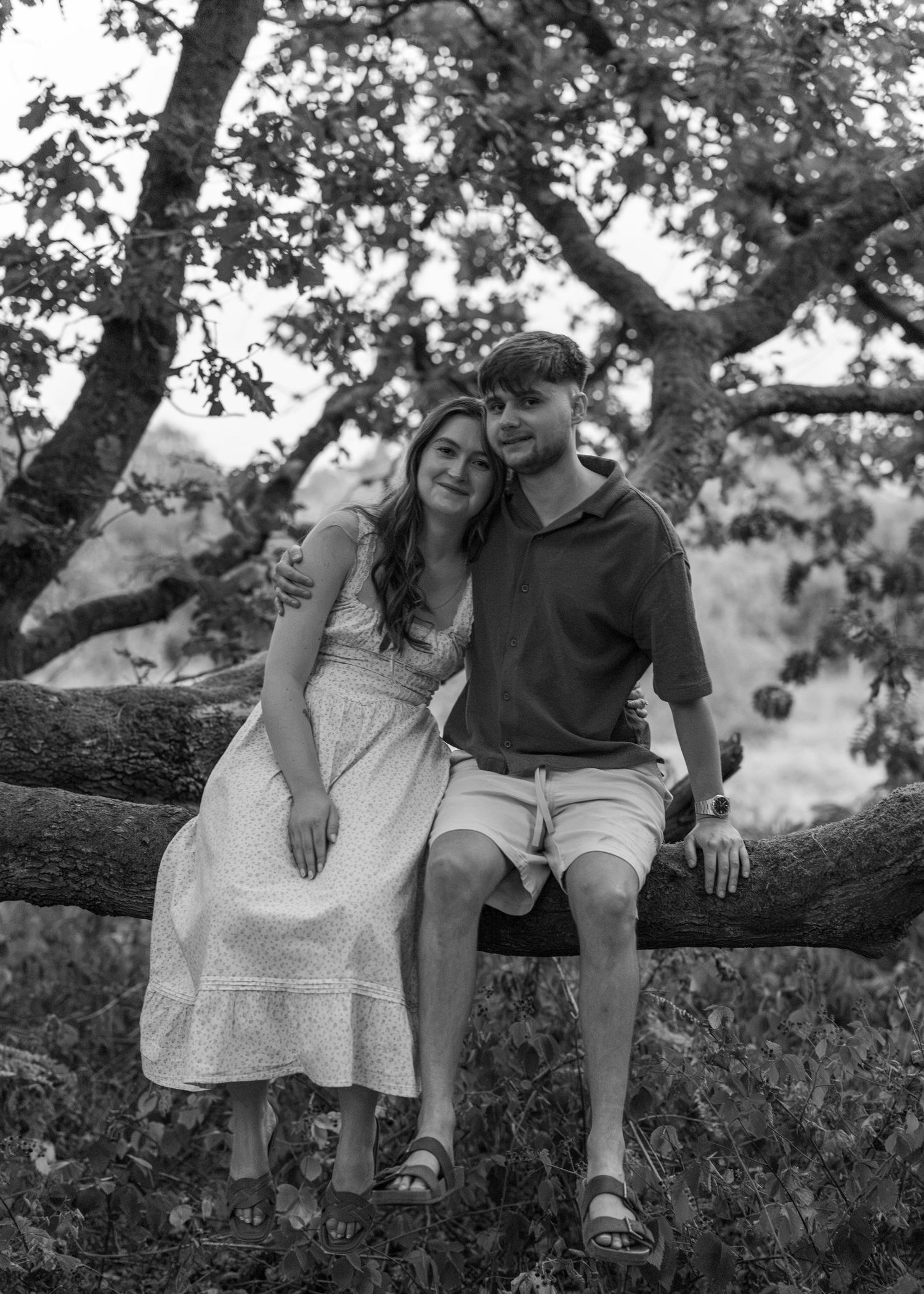 Black and white photo of a young couple sitting on a tree branch outdoors, smiling and embracing in a wooded setting.