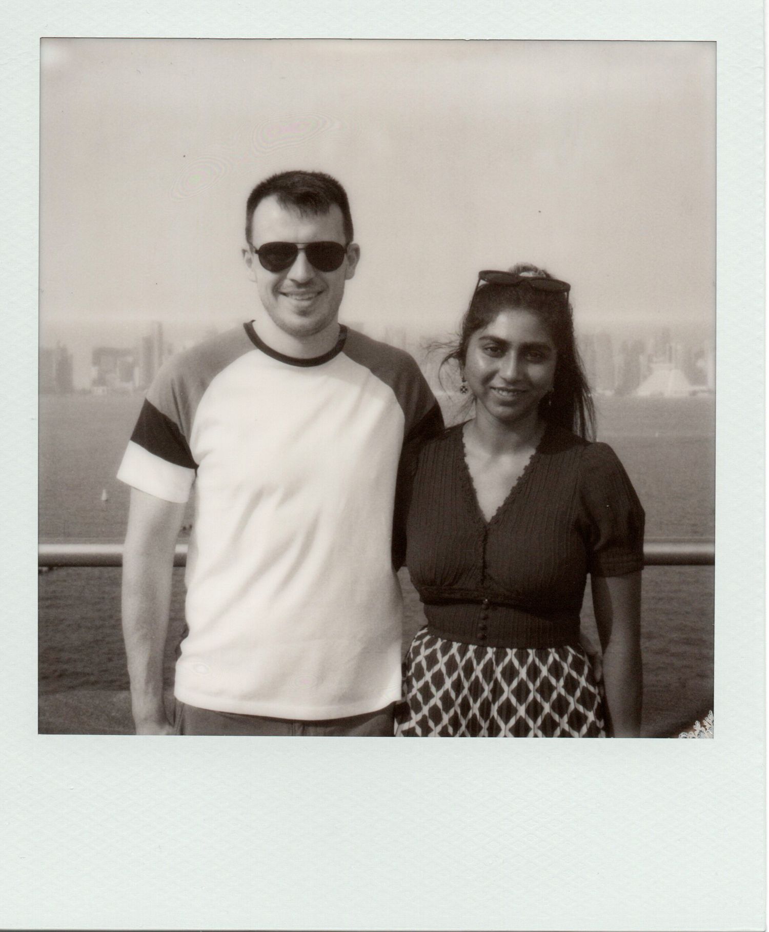 Smiling couple posing together on a waterfront promenade in black and white Polaroid.