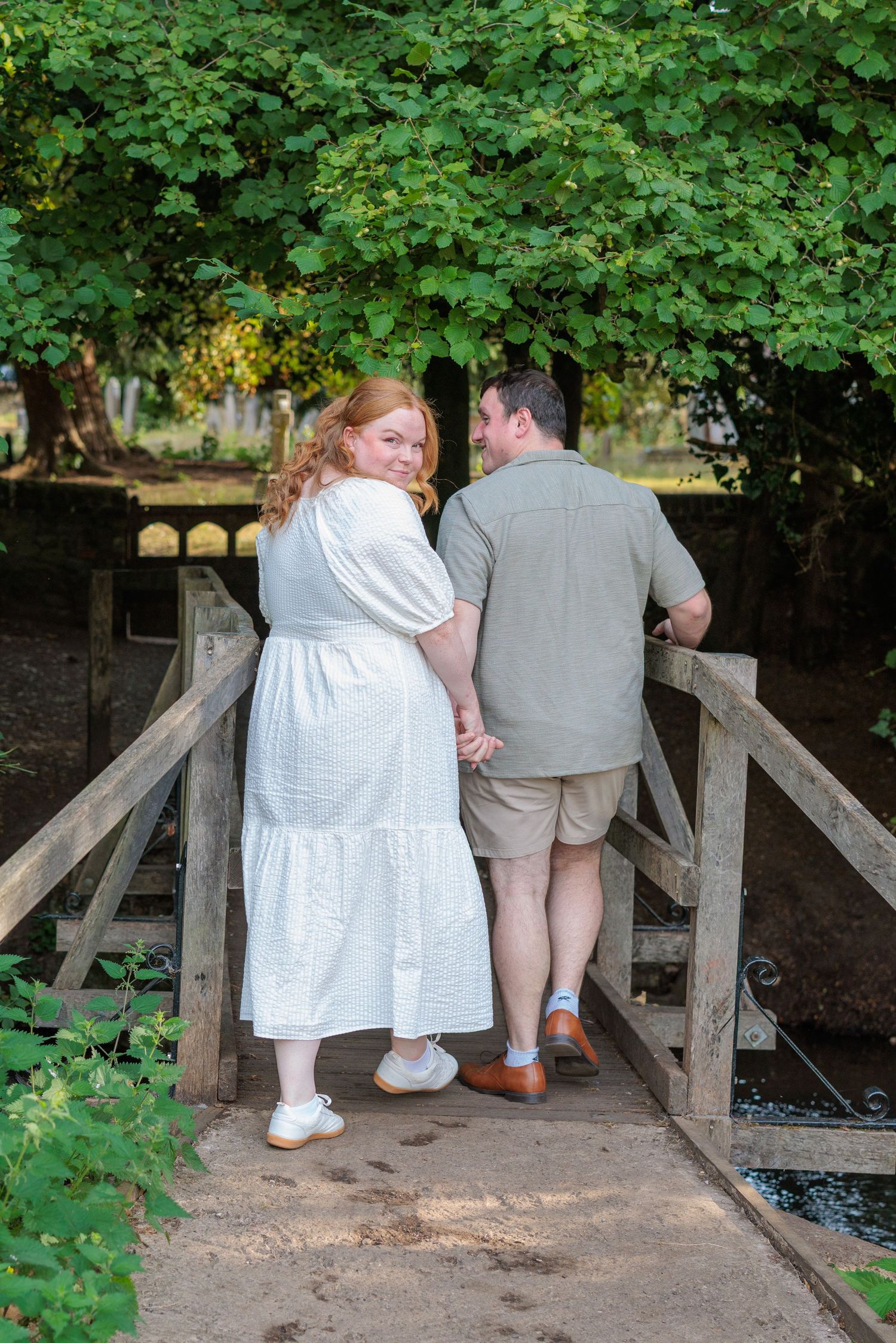 Couple holding hands on a rustic wooden bridge surrounded by lush green trees in a romantic outdoor setting.