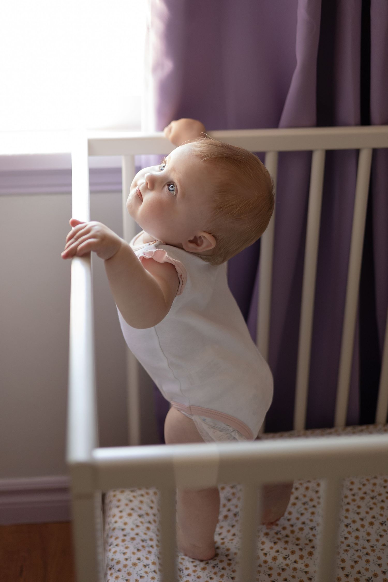 A baby stands in a crib by a window looking up at it's mom.