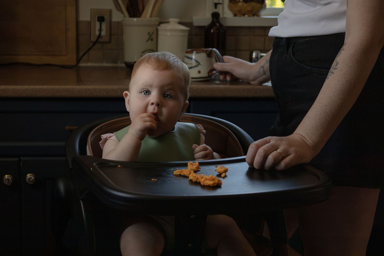 A baby sits in a high chair eating food while looking at the camera. The mom stands beside trying to help.