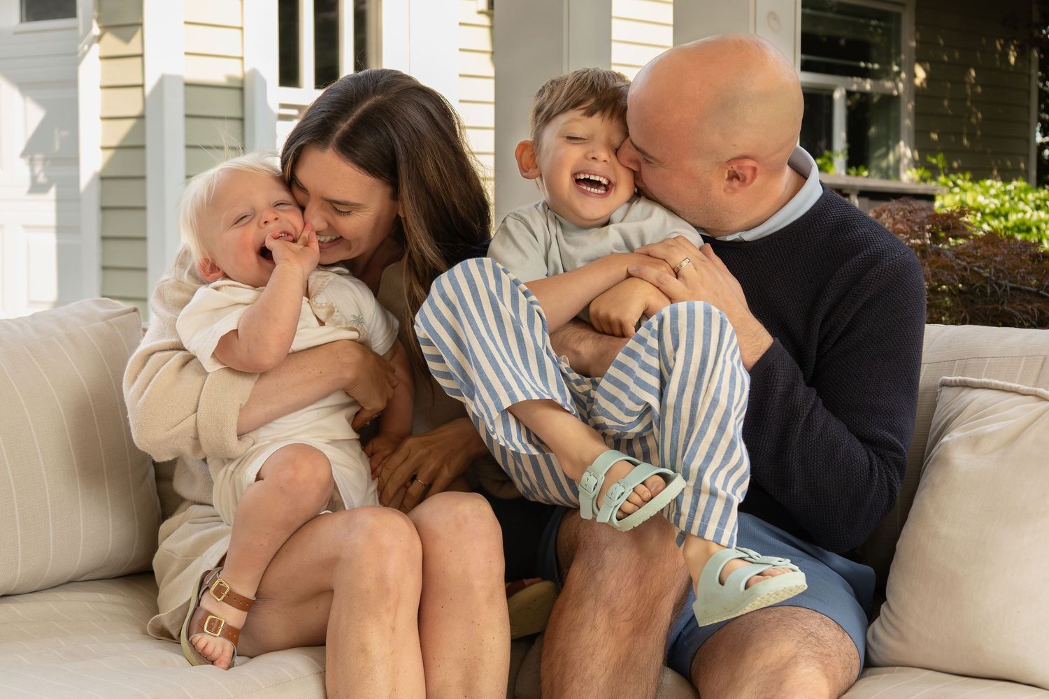 A family with two young boys cuddle and laugh together on a couch in their backyard.