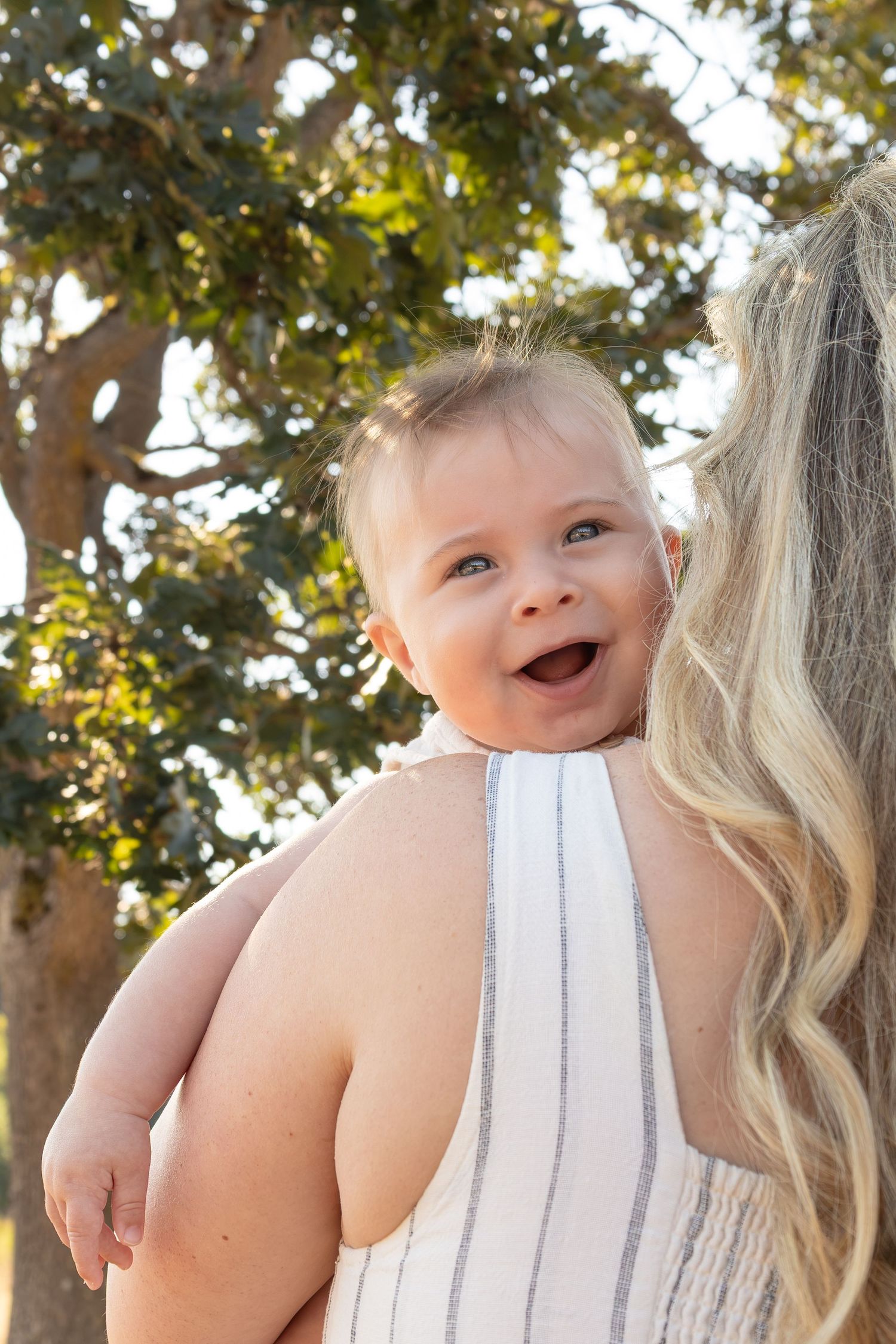A mom holds a smiling baby over her shoulder while standing beneath a tree.