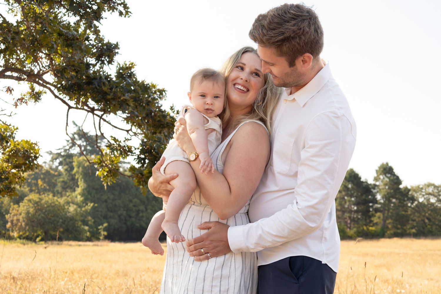 Two parents hold their young baby in a field. The dad hugs the mom from behind while the mom looks up at her husband and smiles.