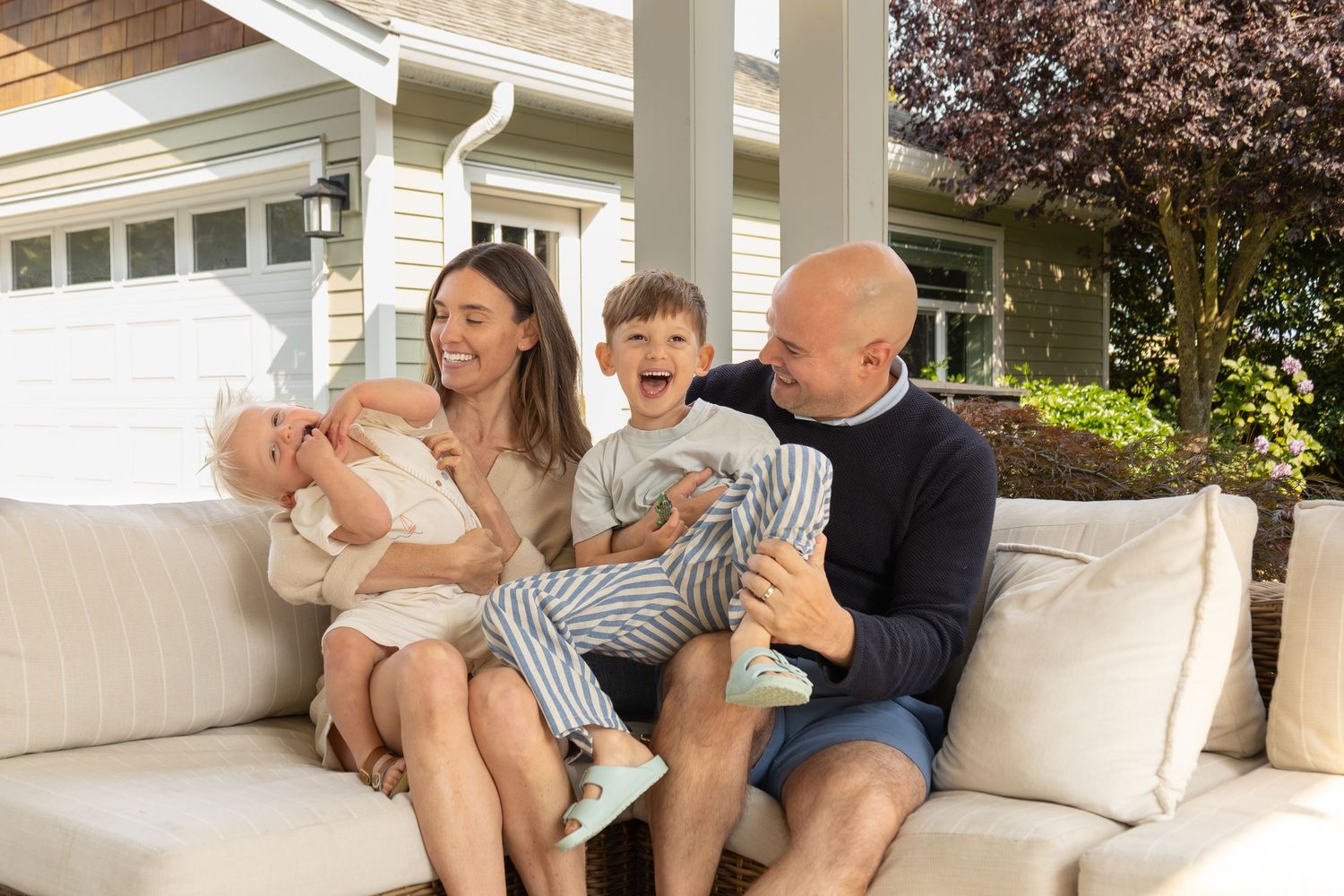 A family with two young boys cuddle and laugh together on a couch in their backyard.
