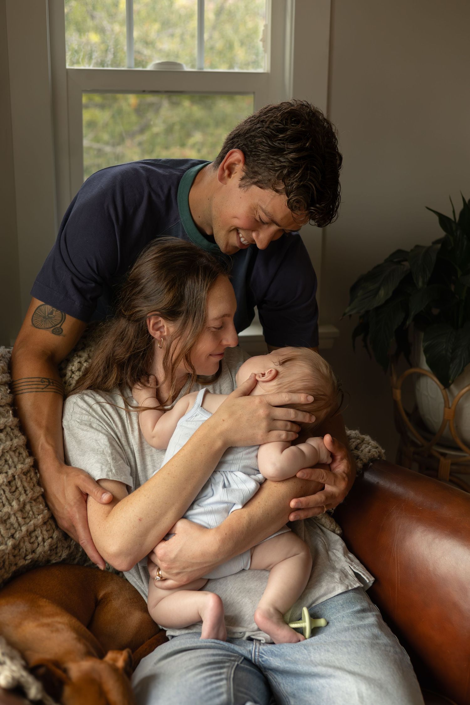 A young mom sits in a chair in a living room with a dog while holding a baby who is standing on her lap. The dad stands behind watching.