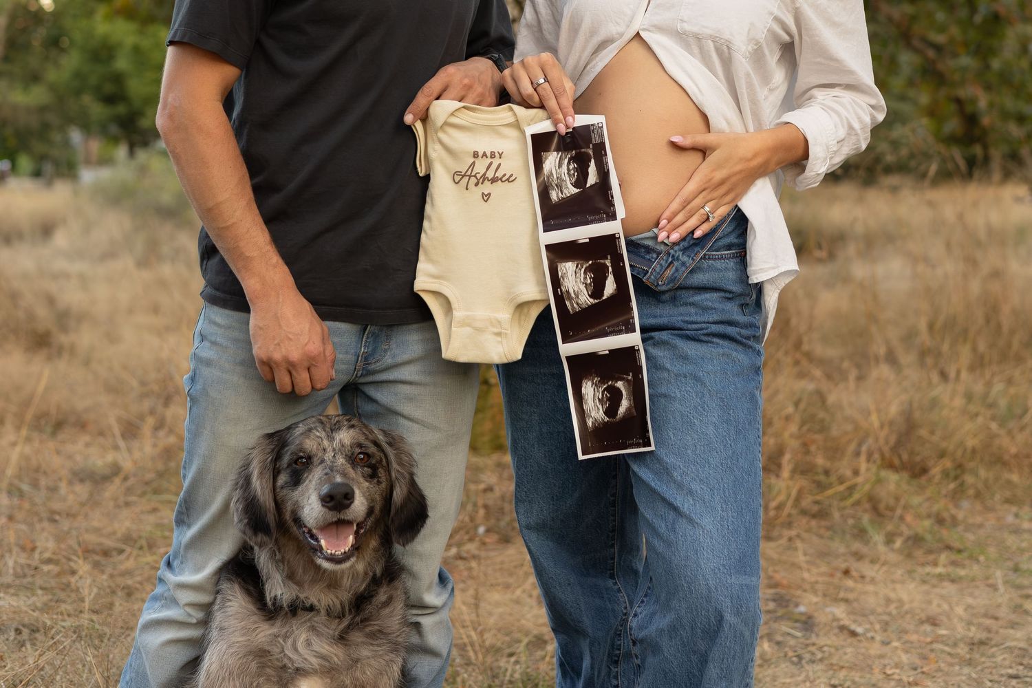 A couple stands together with their dog. They hold a baby onesie and a sonogram and the women holds her belly to announce her pregnancy.