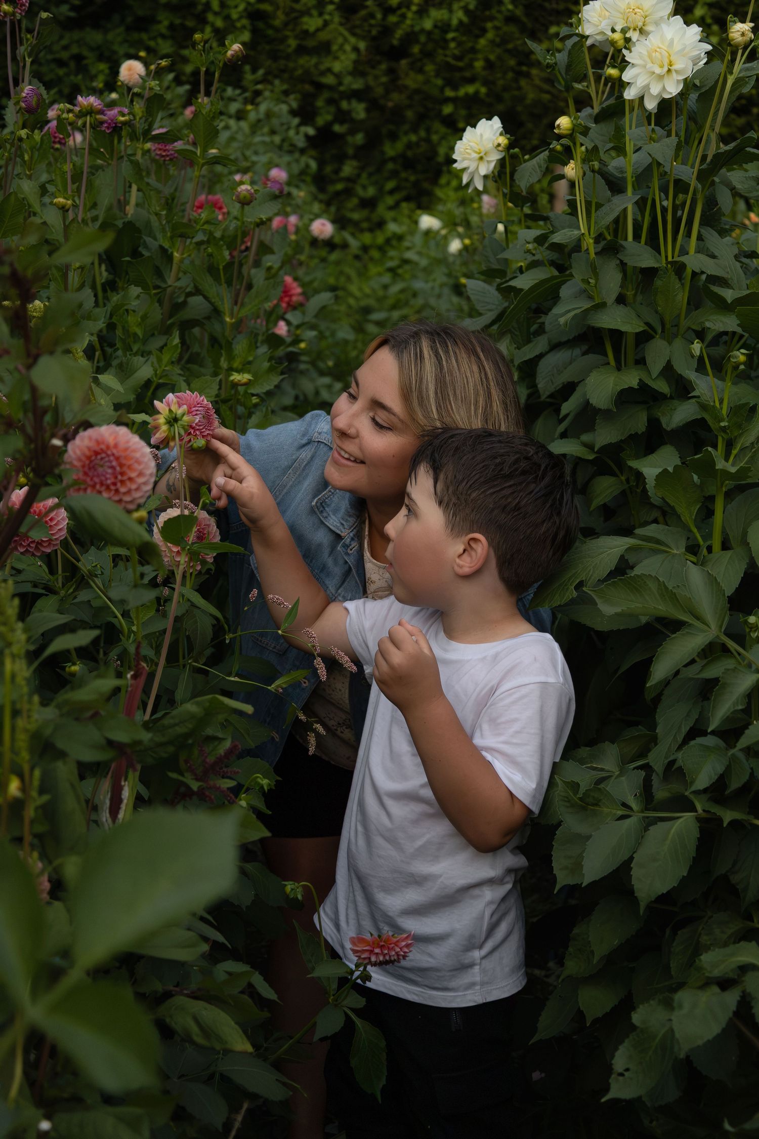 A mom and her young son explore a flower field. The boy points to a flower and asks his mom a question. They smile and look at it together.